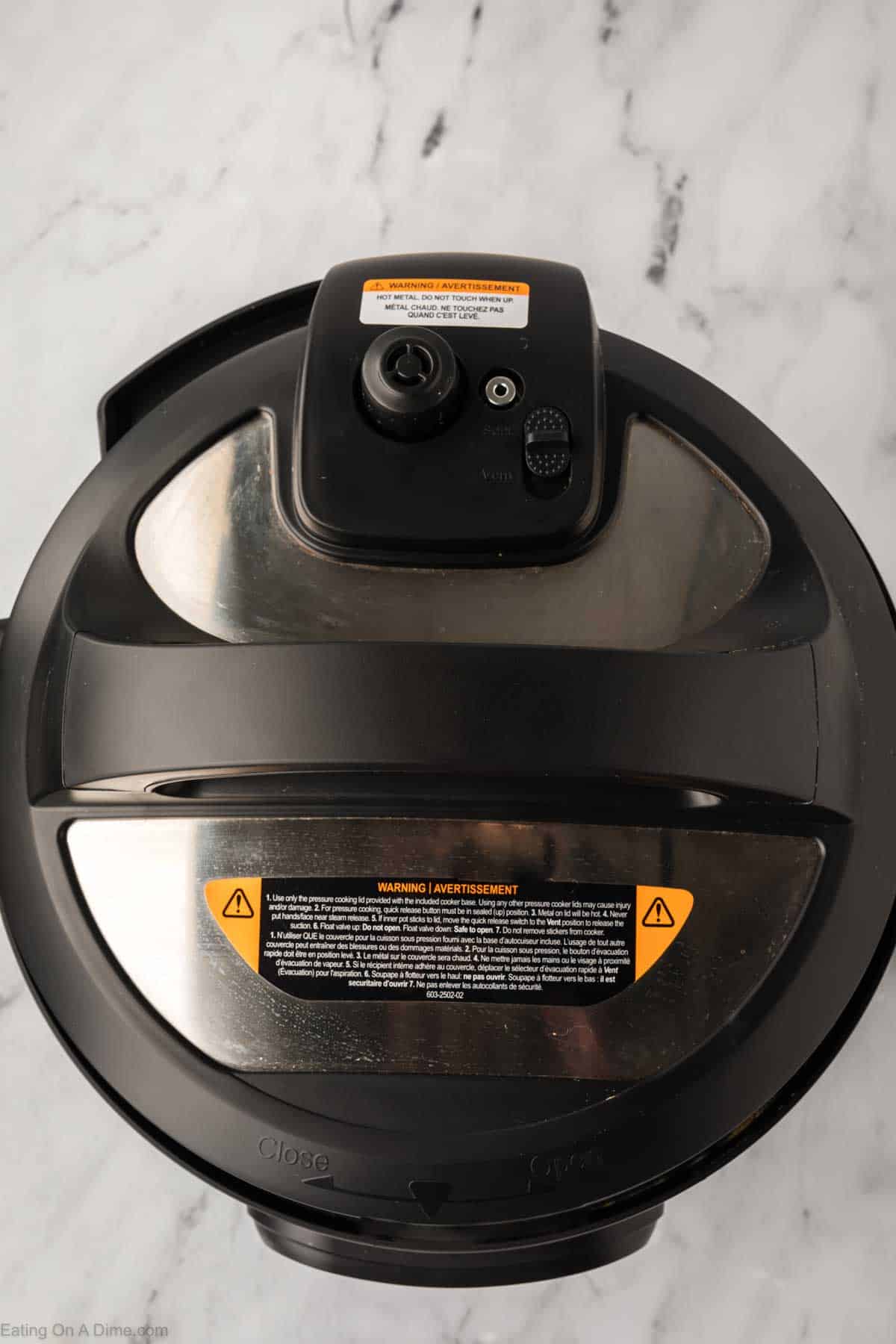 Top view of a black and silver Instant Pot lid on a marble surface. The lid, perfect for cooking Whole Chicken, features warning labels and a pressure release valve.