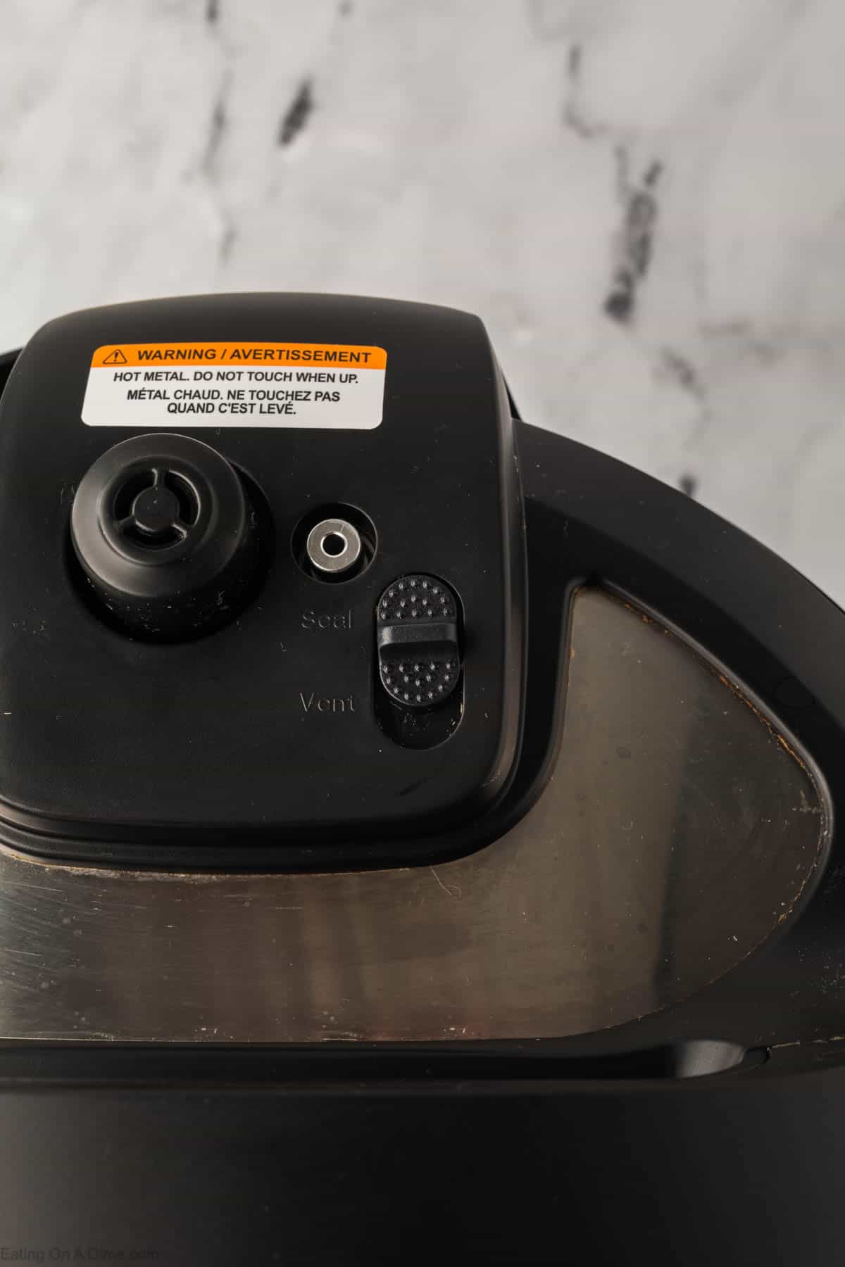 Close-up of a pressure cooker lid, perfect for preparing an Instant Pot whole chicken, featuring a steam release valve with "Seal" and "Vent" options. The sleek black surface showcases a safety warning label on top, all set against a marble countertop background.