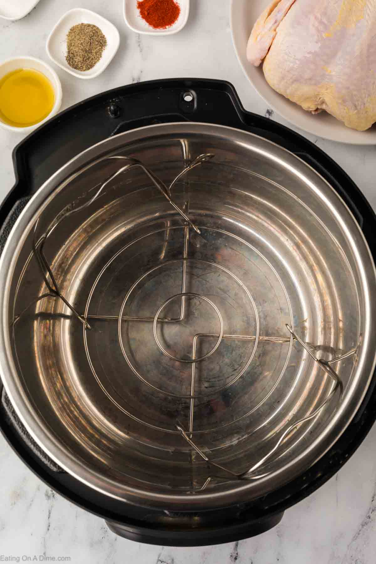 An open Instant Pot with a metal trivet inside rests on a marble countertop. Surrounding it are bowls filled with black pepper, paprika, and salt, ready to season the whole chicken that's partially visible on a white plate. Perfect setup to cook an Instant Pot Whole Chicken masterpiece.