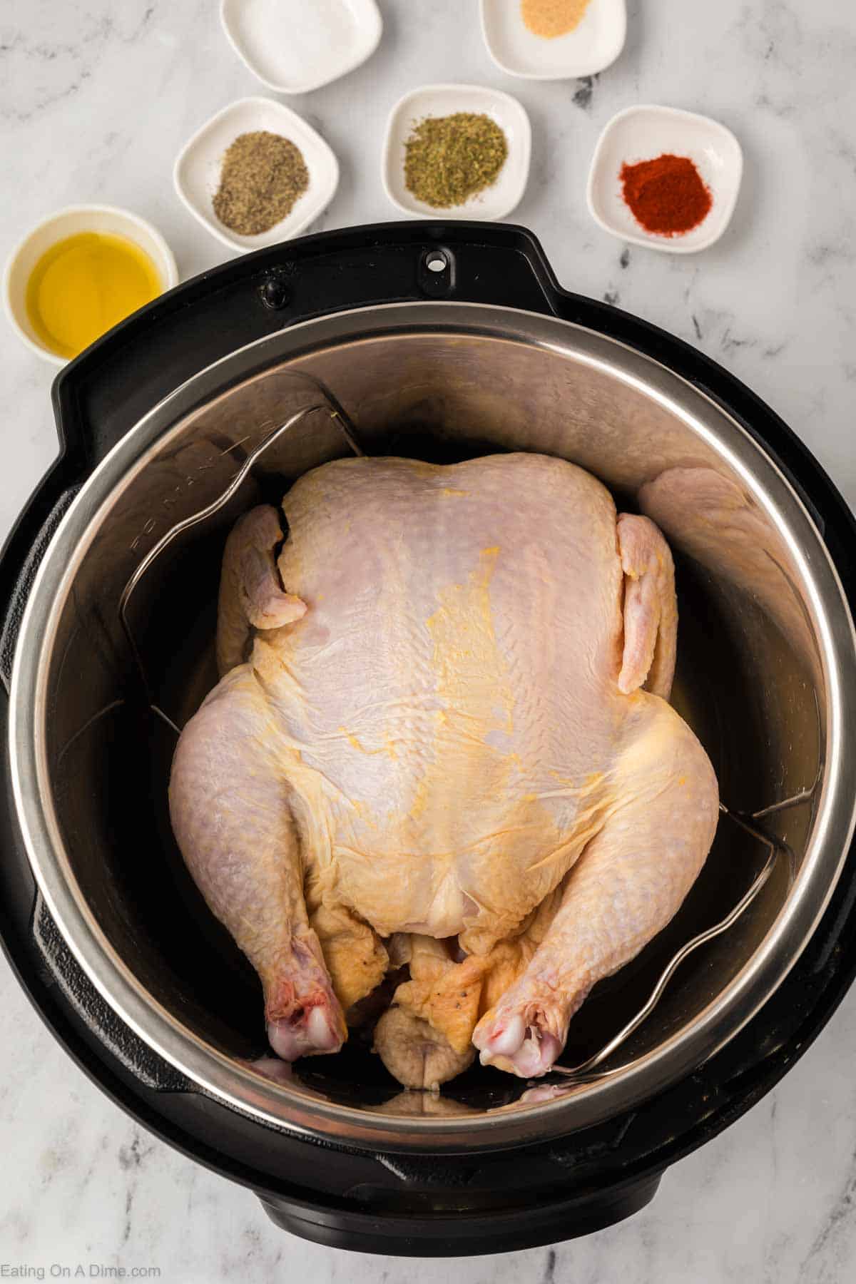 A raw whole chicken inside an Instant Pot sits ready to be transformed. Surrounding the black pot are small bowls filled with a variety of spices and seasonings, all resting on a sleek marble surface.