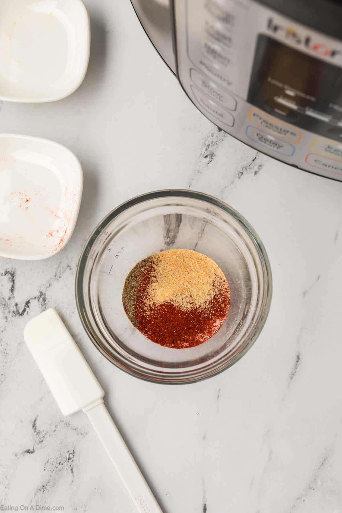 A glass bowl on the marble countertop holds a blend of powdered spices, perfect for seasoning your Instant Pot whole chicken. Nearby, a small white spatula and two empty dishes await their task. In the top right corner, part of a kitchen appliance peeks into view.