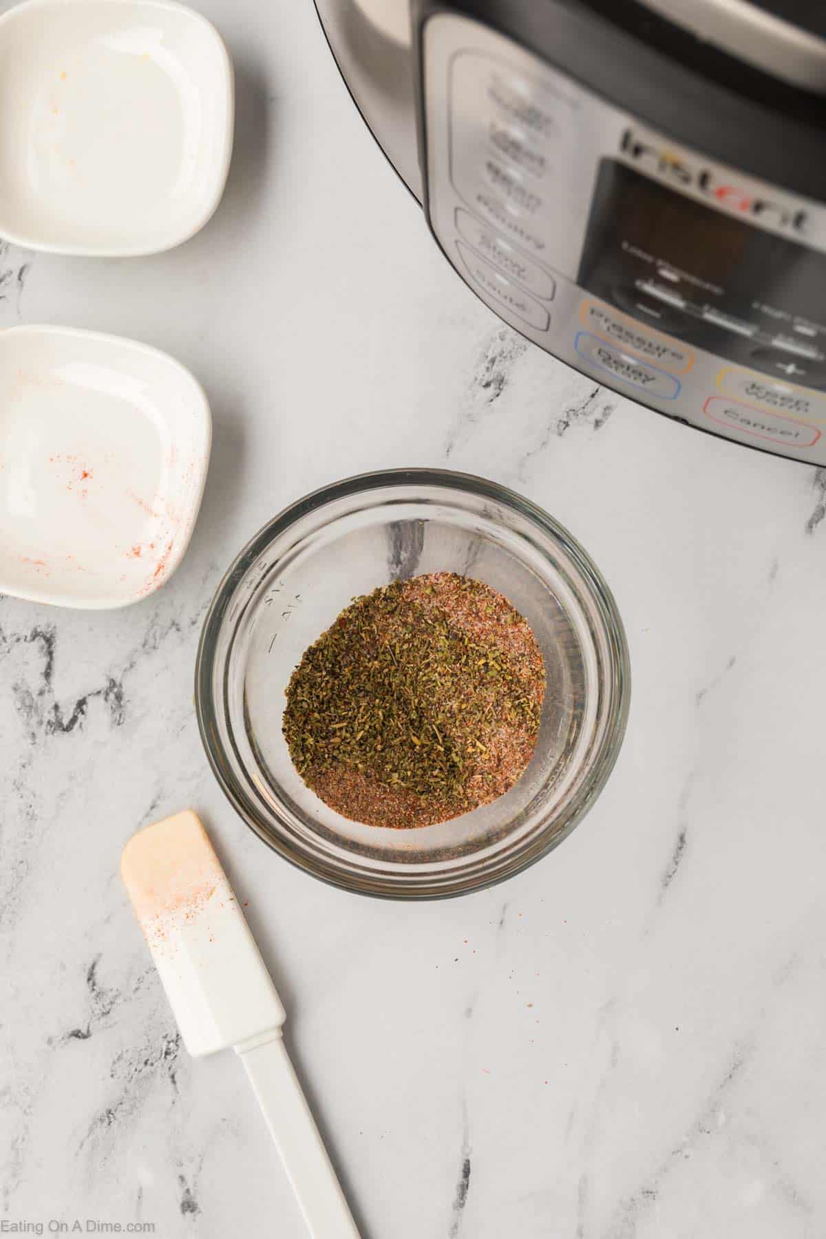 A small glass bowl containing spices sits on a marble countertop, next to an empty white dish with some spice residue. A white spatula is beside the bowl, while part of an Instant Pot—perfect for preparing a whole chicken—is visible in the top right corner.