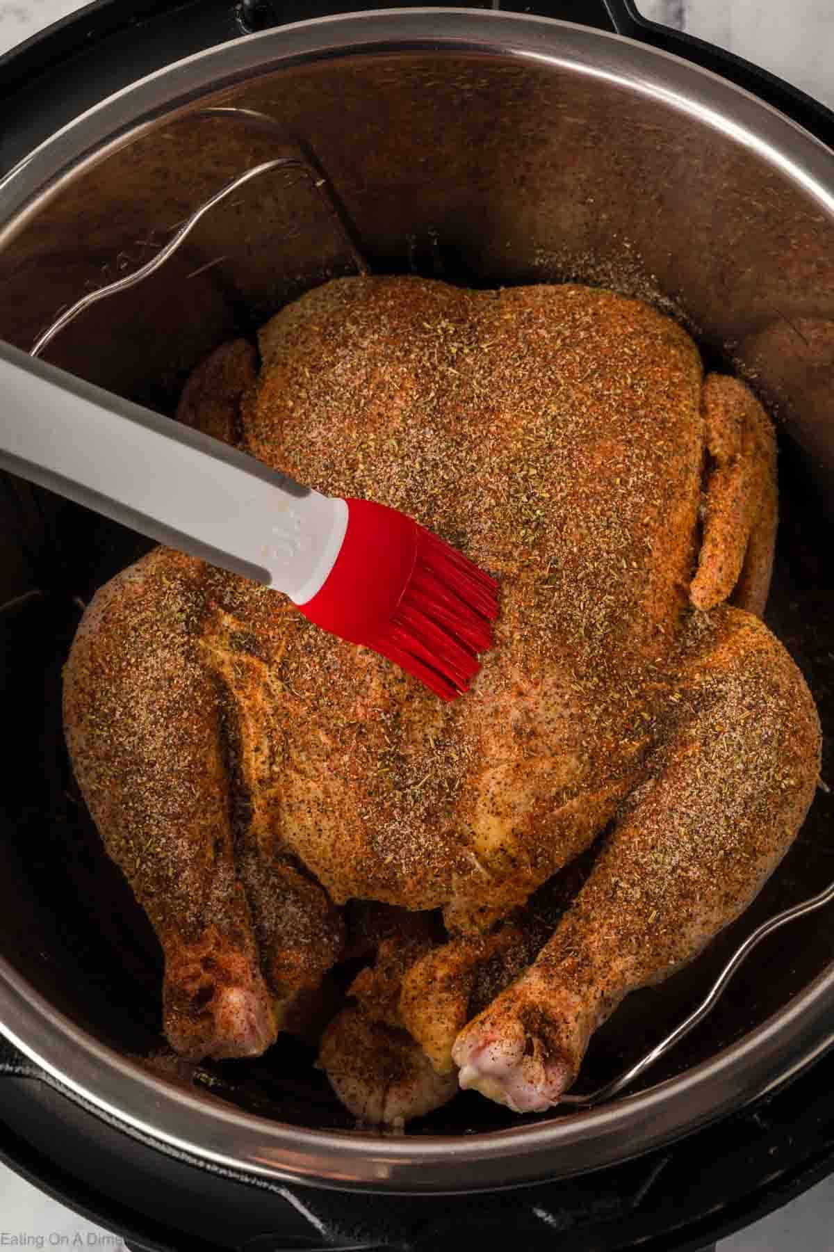 An Instant Pot Whole Chicken sits ready for its culinary transformation. A red silicone basting brush glides marinade over the chicken, which is generously covered with herbs and spices, poised for perfect cooking.