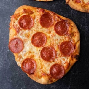 A homemade flatbread pizza with a crispy crust and melted cheese rests on a dark surface. The pizza is topped with evenly spaced slices of pepperoni, making it an irresistible treat.