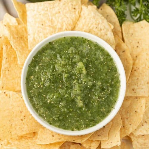 A bowl of salsa verde de tomatillo sits in the center of a plate surrounded by crispy, golden tortilla chips. The salsa has a textured, slightly chunky appearance, and some parsley adds a touch of freshness in the background.