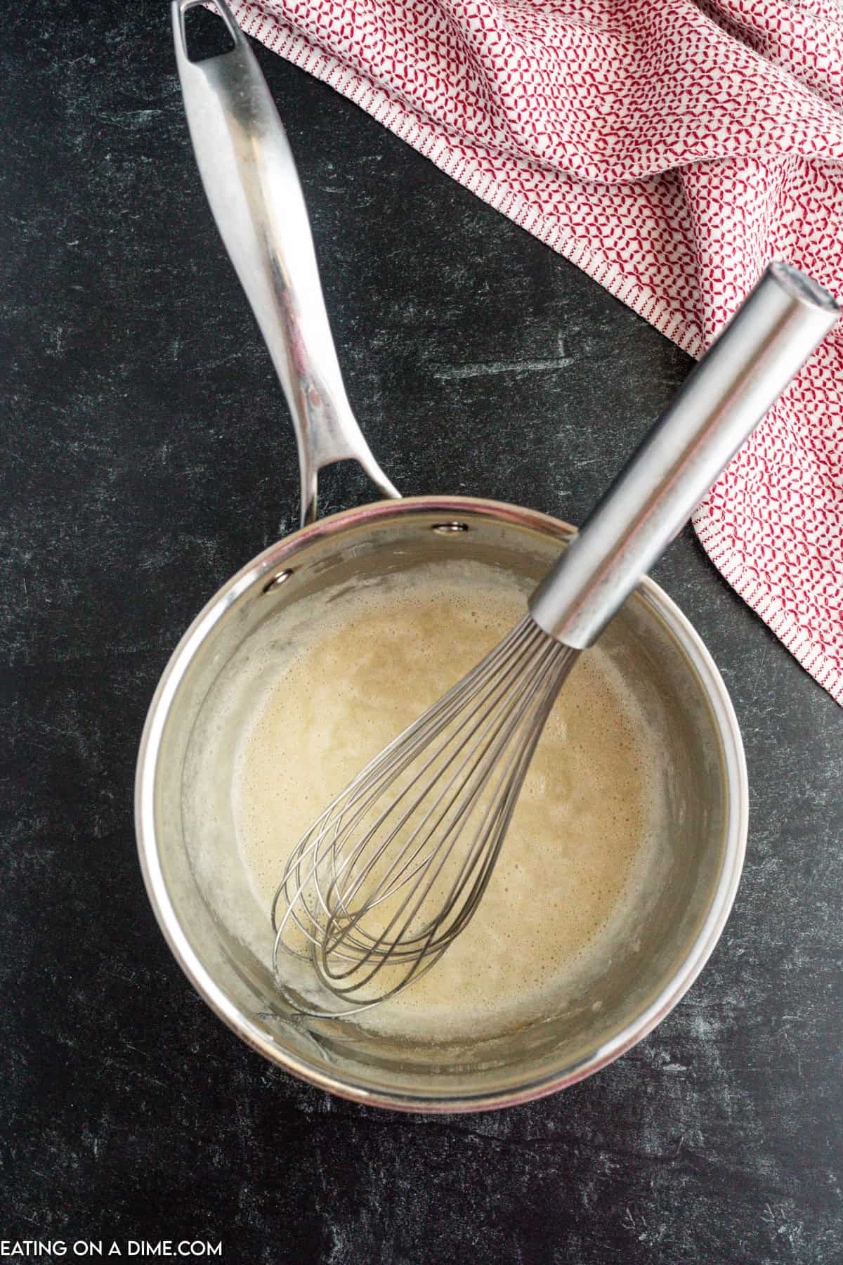 A metal saucepan with a whisk inside it sits on a dark countertop, containing a light brown homemade nacho cheese sauce. A red and white patterned cloth is partially visible near the top edge of the image. The bottom left corner has the text "EATING ON A DIME.COM".