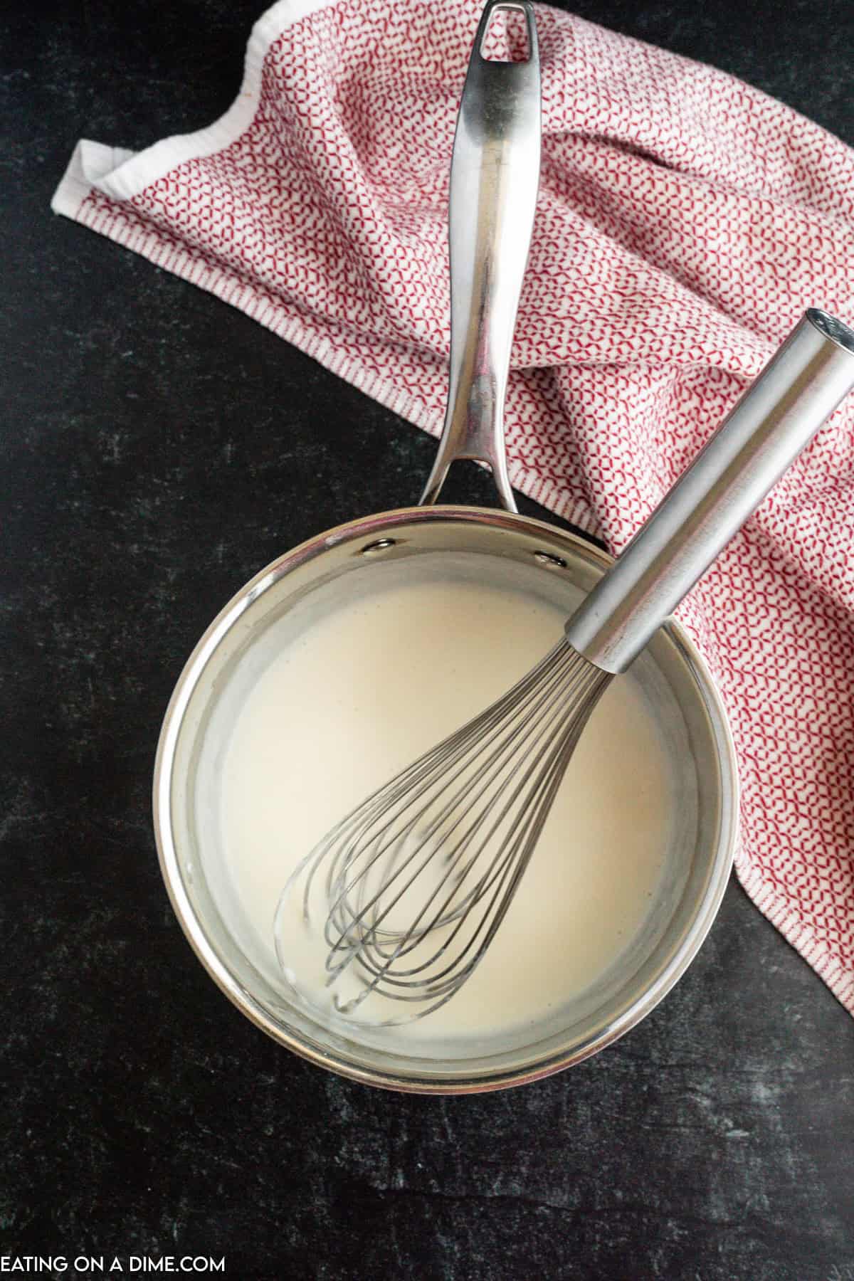 A stainless steel saucepan filled with creamy homemade nacho cheese sauce sits on a black countertop. A metal whisk is inside the saucepan, and a red and white patterned cloth is draped in the background.