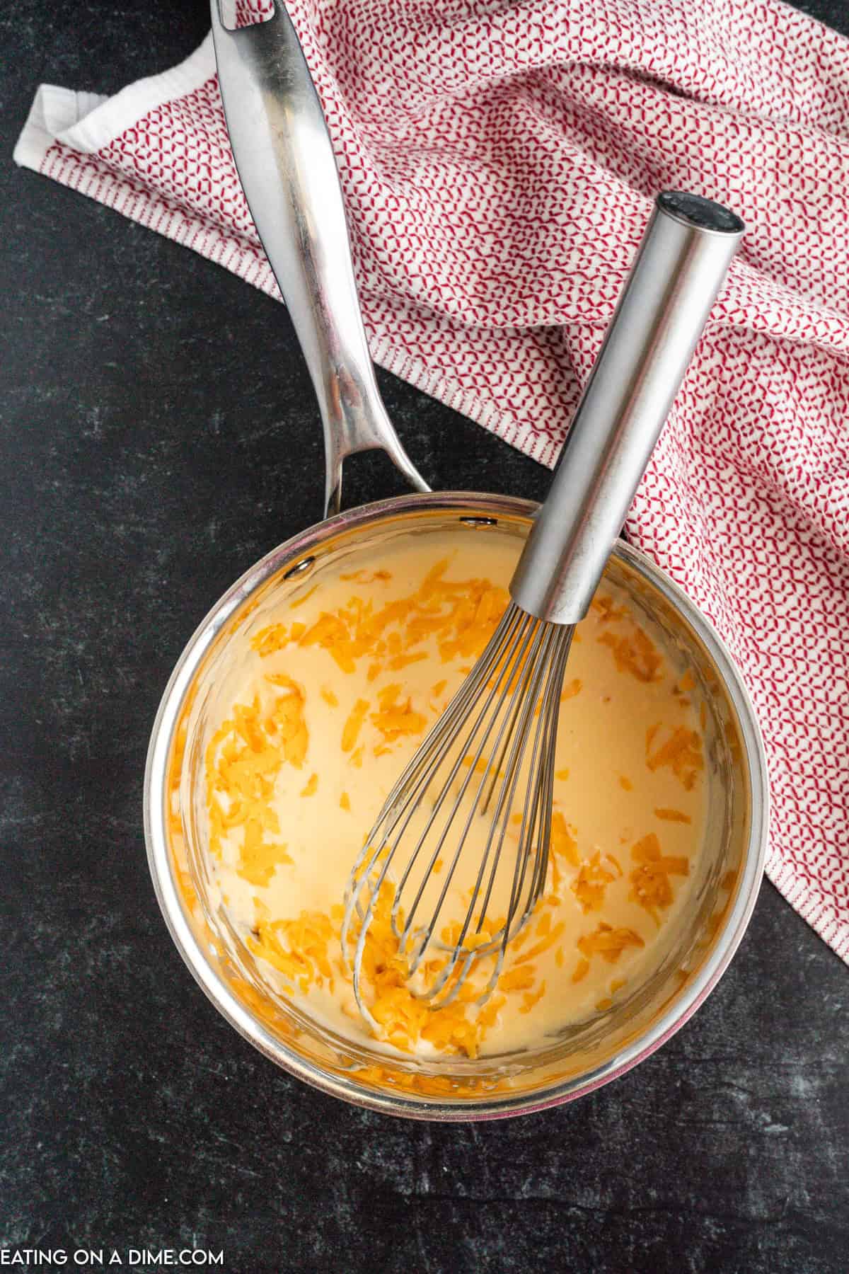 A saucepan filled with homemade nacho cheese sauce, partially mixed with shredded cheese, sits on a dark countertop. A whisk rests in the saucepan. A red and white patterned kitchen towel is placed beside it.