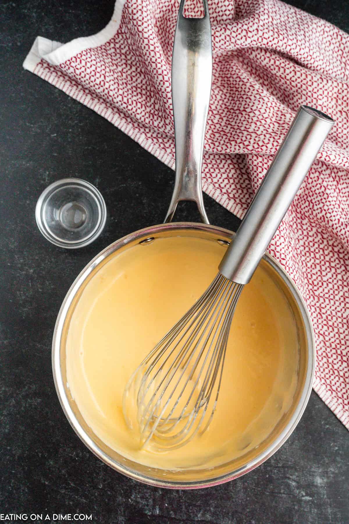 A stainless steel saucepan filled with a creamy yellow homemade nacho cheese sauce, mixed by a whisk also in the pan. The saucepan is on a dark countertop next to a small glass bowl and a red and white patterned dish towel. The edges of the towel are slightly draped over the countertop.