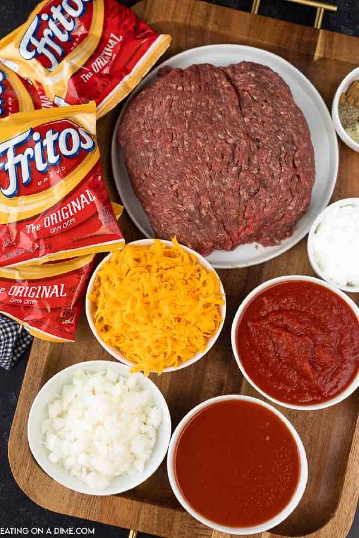 Ingredients for a classic chili dish, reminiscent of a walking taco, are arranged on a wooden board. Visible are ground meat, shredded cheddar cheese, tomato sauce, diced onions, and bags of Fritos corn chips. Seasonings and bowls of additional sauce complete the setup.