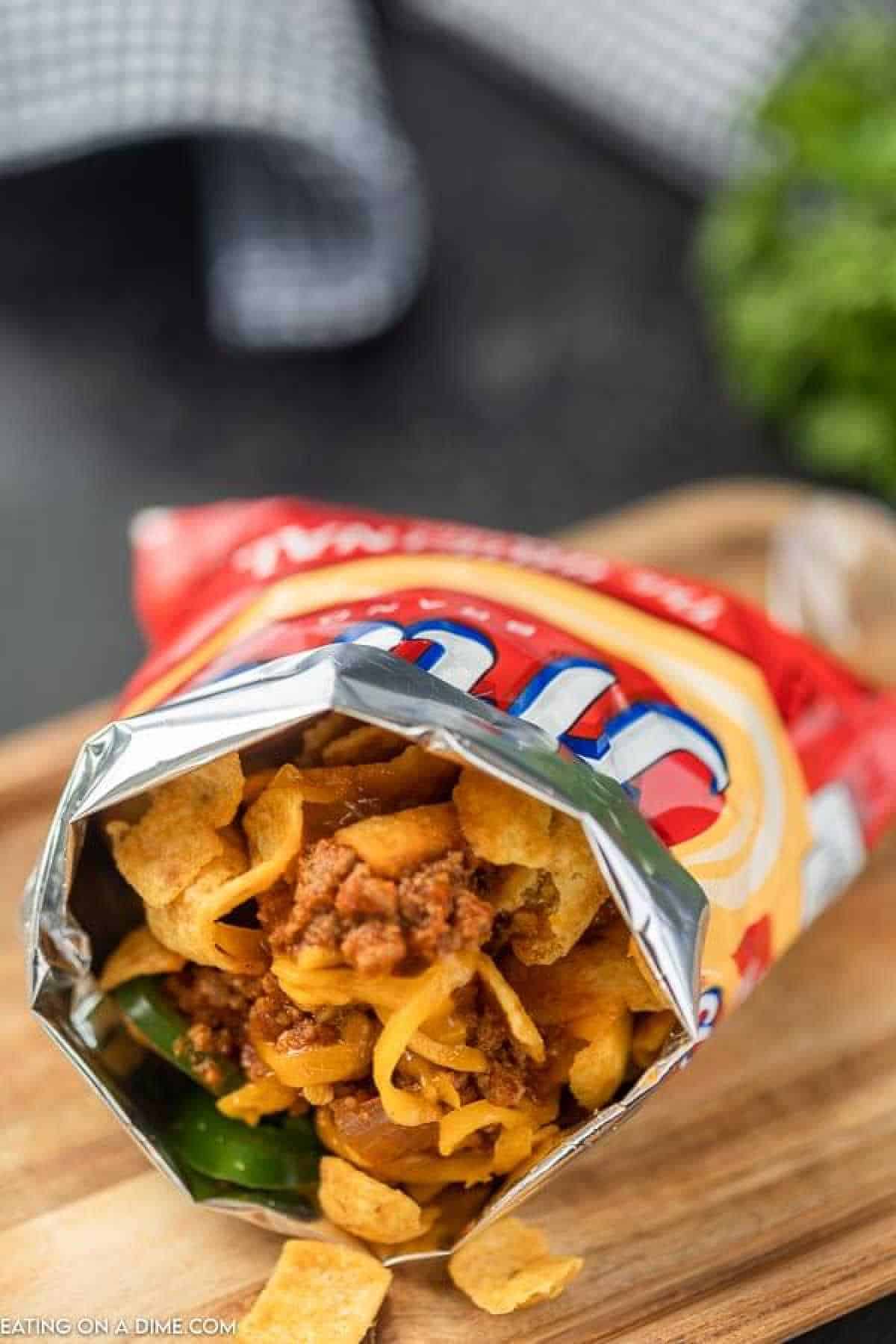 Open bag of Fritos filled with chili, cheese, and sliced jalapeños, creating a delectable walking taco sensation. Placed on a wooden surface, the scene is complemented by a checkered cloth and some greens in the blurred background.