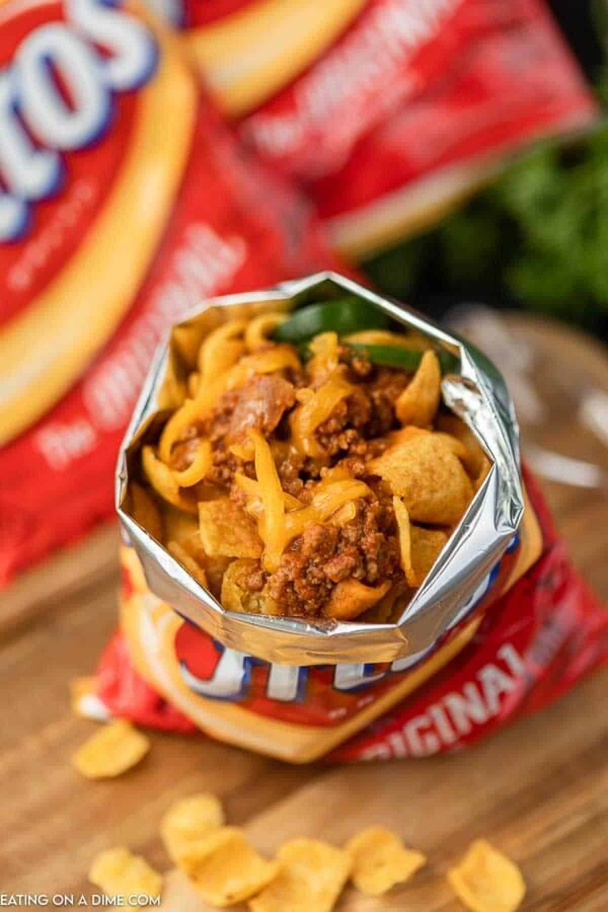 An open bag of Fritos brimming with chili and topped with melted cheese rests on a wooden surface, capturing the essence of a walking taco. In the background, an unopened Fritos bag awaits its turn in this delicious frito chili pie experience.