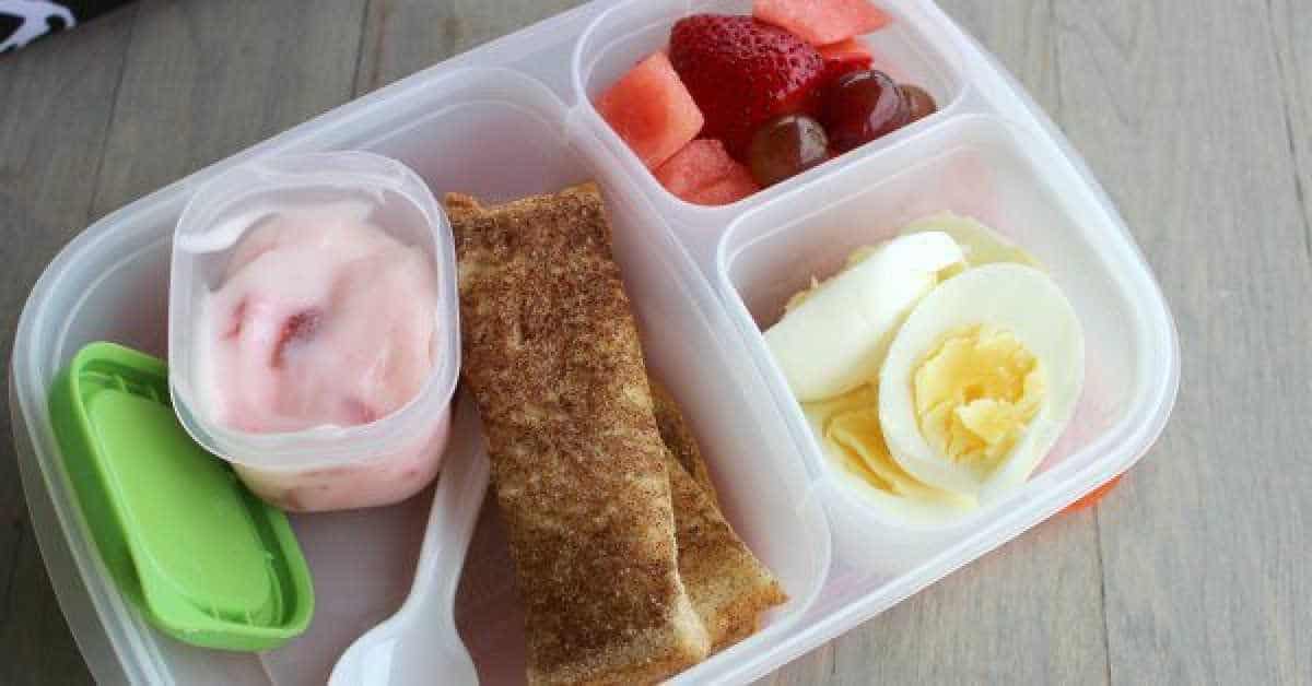 A divided lunch container features yogurt and strawberries, cinnamon toast, assorted fruits like grapes and watermelon, and a hard-boiled egg for simple lunch ideas. A green sauce container and white plastic spoon complete the set.