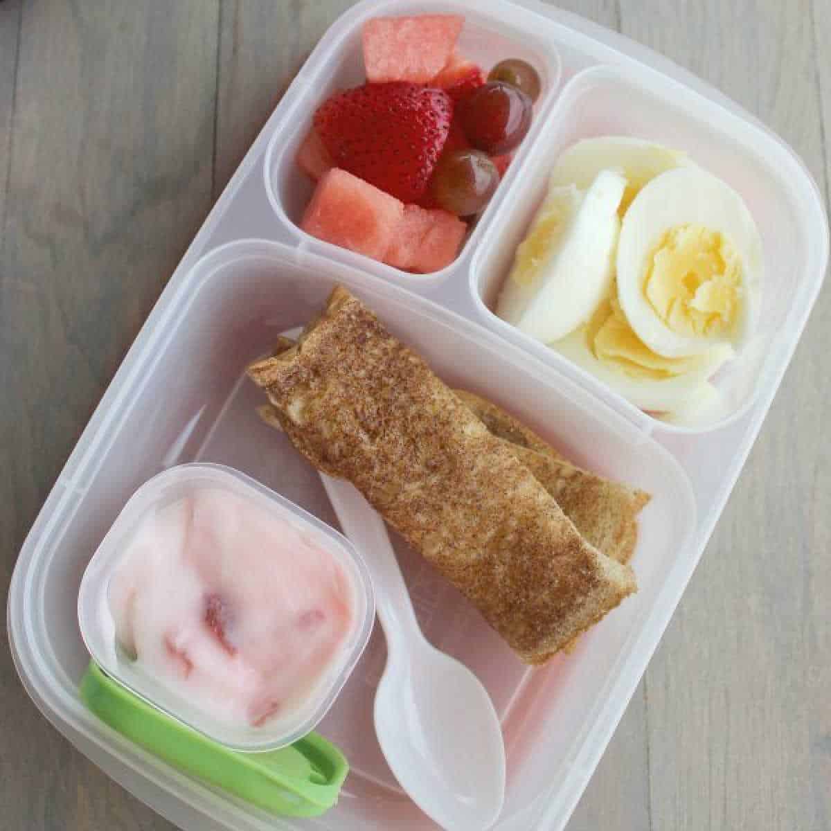 A bento box offers hard boiled egg lunch ideas with compartments of sliced eggs, mixed fruits like strawberries and grapes, yogurt speckled with fruit bits, and rolled flatbread. Inside the box, a spoon awaits to assist your flavorful journey.