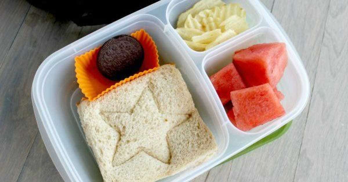 A lunchbox features a cookie cutter sandwich with a star pattern, neatly placed in its compartment. Accompanying it are watermelon cubes, crispy potato chips, and a chocolate sandwich cookie in an orange wrapper. The container rests on a light wood surface.