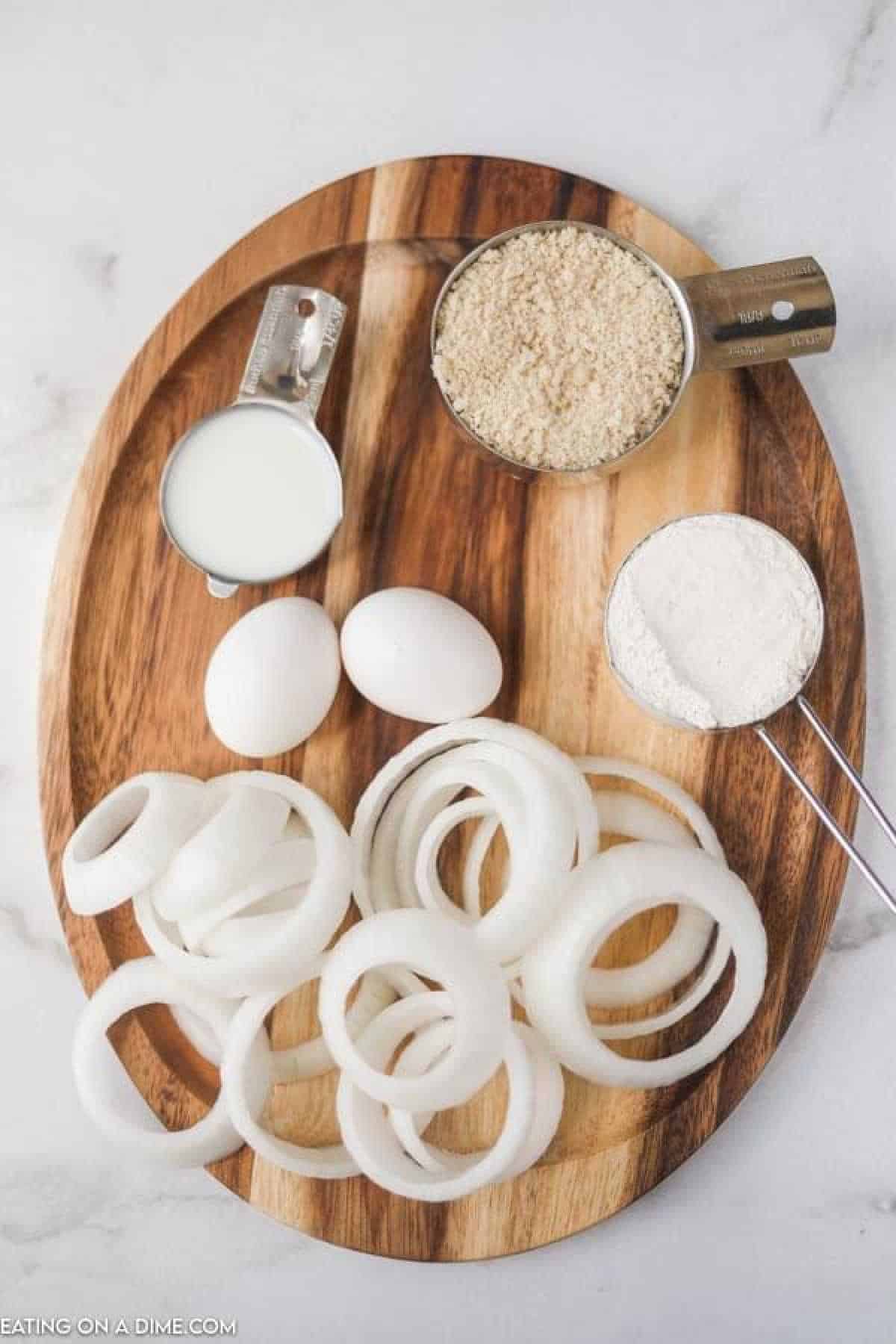 Ingredients for making delicious fried onion rings are arranged on a wooden board: fresh onion slices, two eggs, a cup of milk, breadcrumbs, and flour.