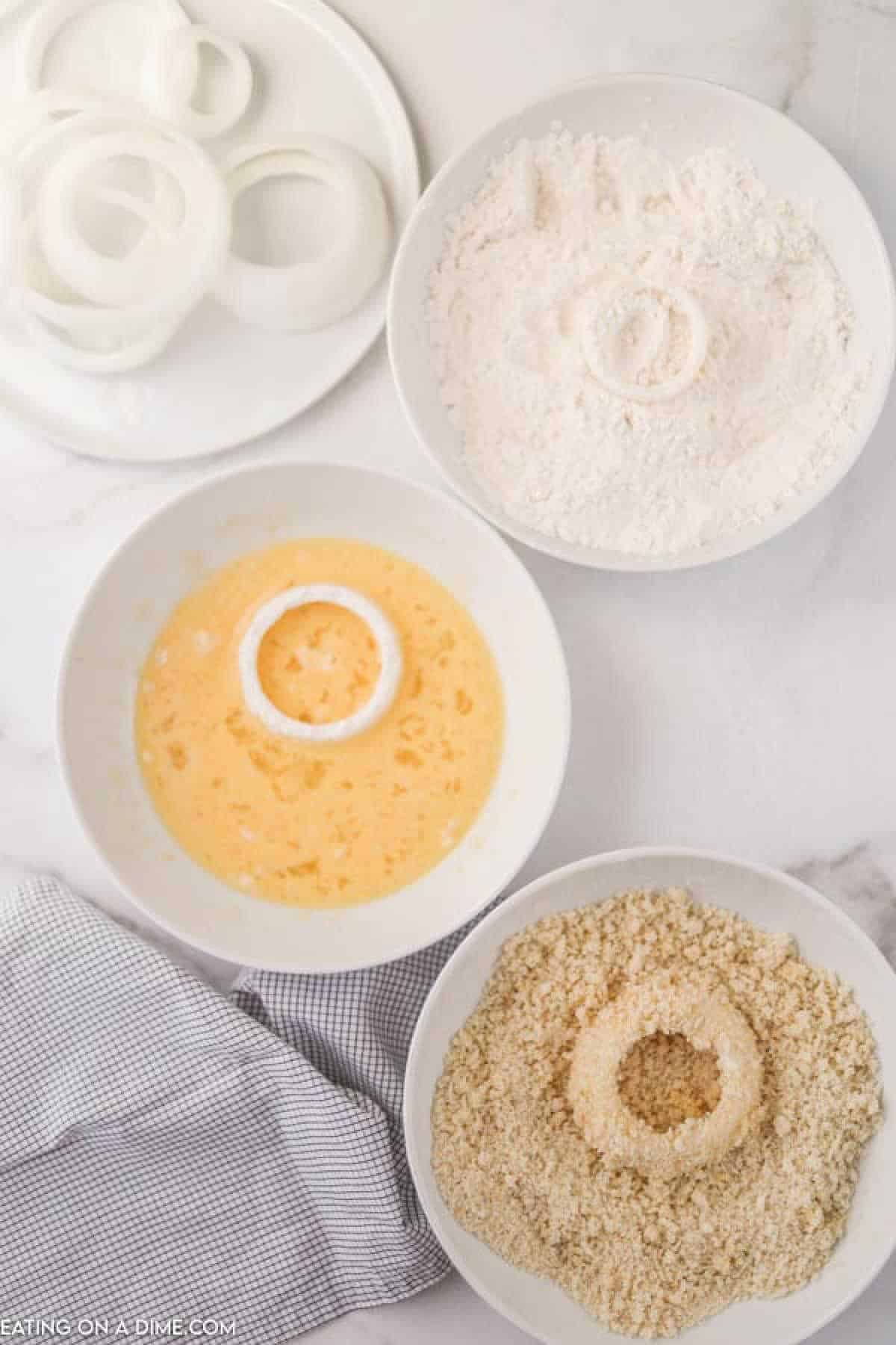 Three bowls showcase the fried onion ring coating process: sliced onion rings rest on a plate, flour fills one bowl, egg mixture occupies another, and breadcrumbs complete the set. A dish towel lies nearby on a light-colored surface.