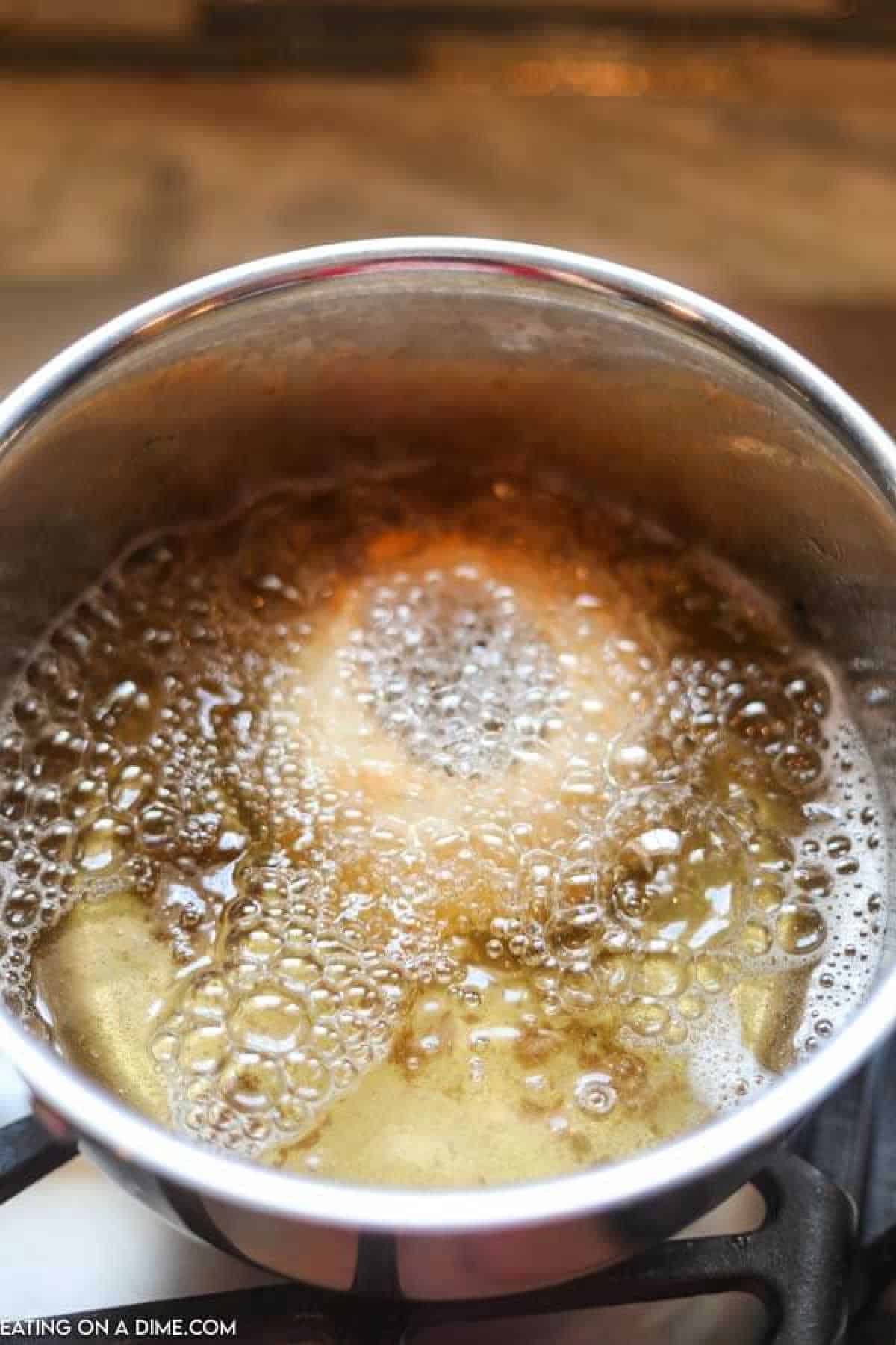 A close-up of a pot filled with bubbling caramel sauce on a stovetop, reminiscent of the golden hues found on crispy fried onion rings. The sauce is foamy and golden brown, indicating it is being heated to the boiling point.