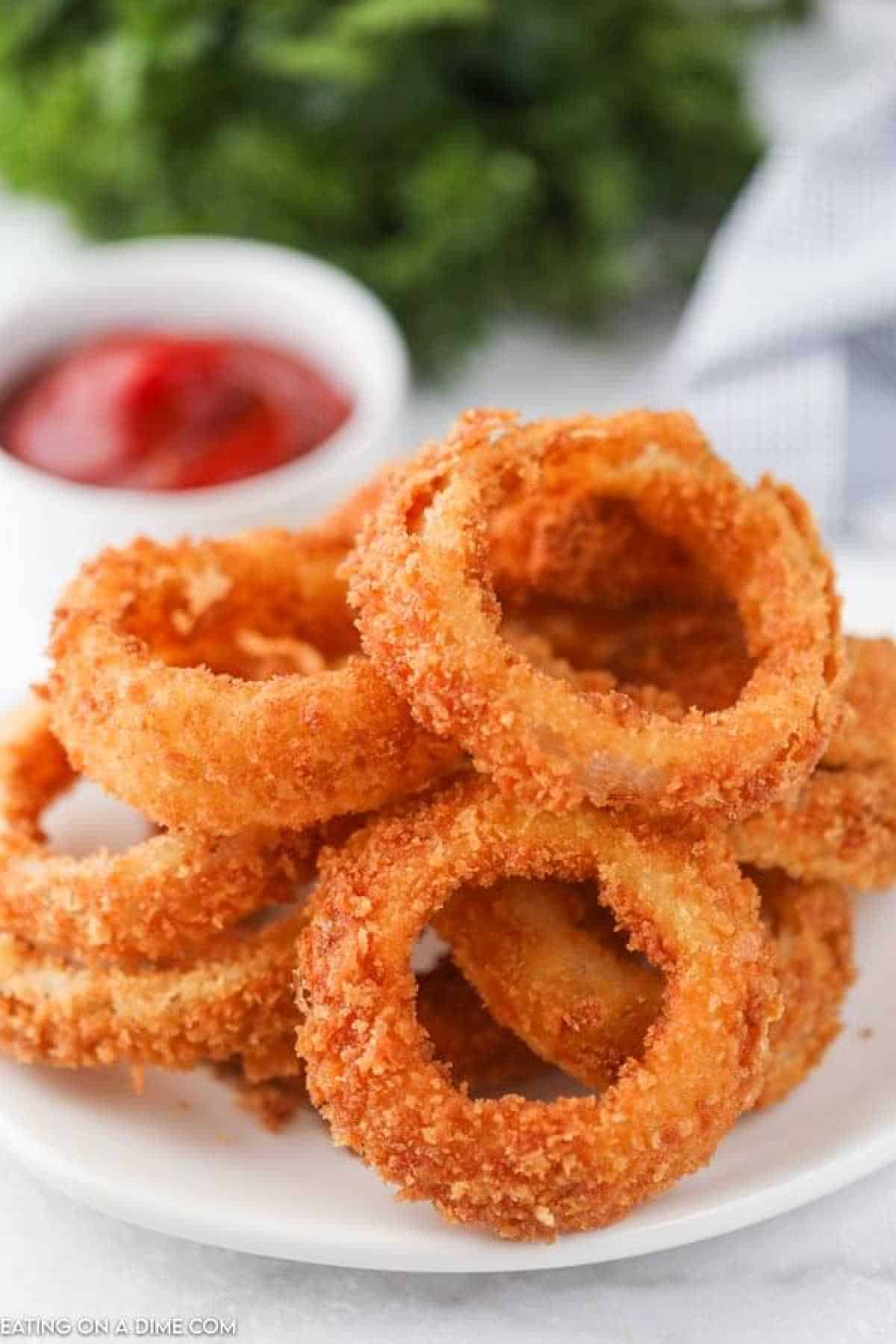 A plate of crispy, fried onion rings stacked neatly, with a small dish of red dipping sauce in the background on a white surface.