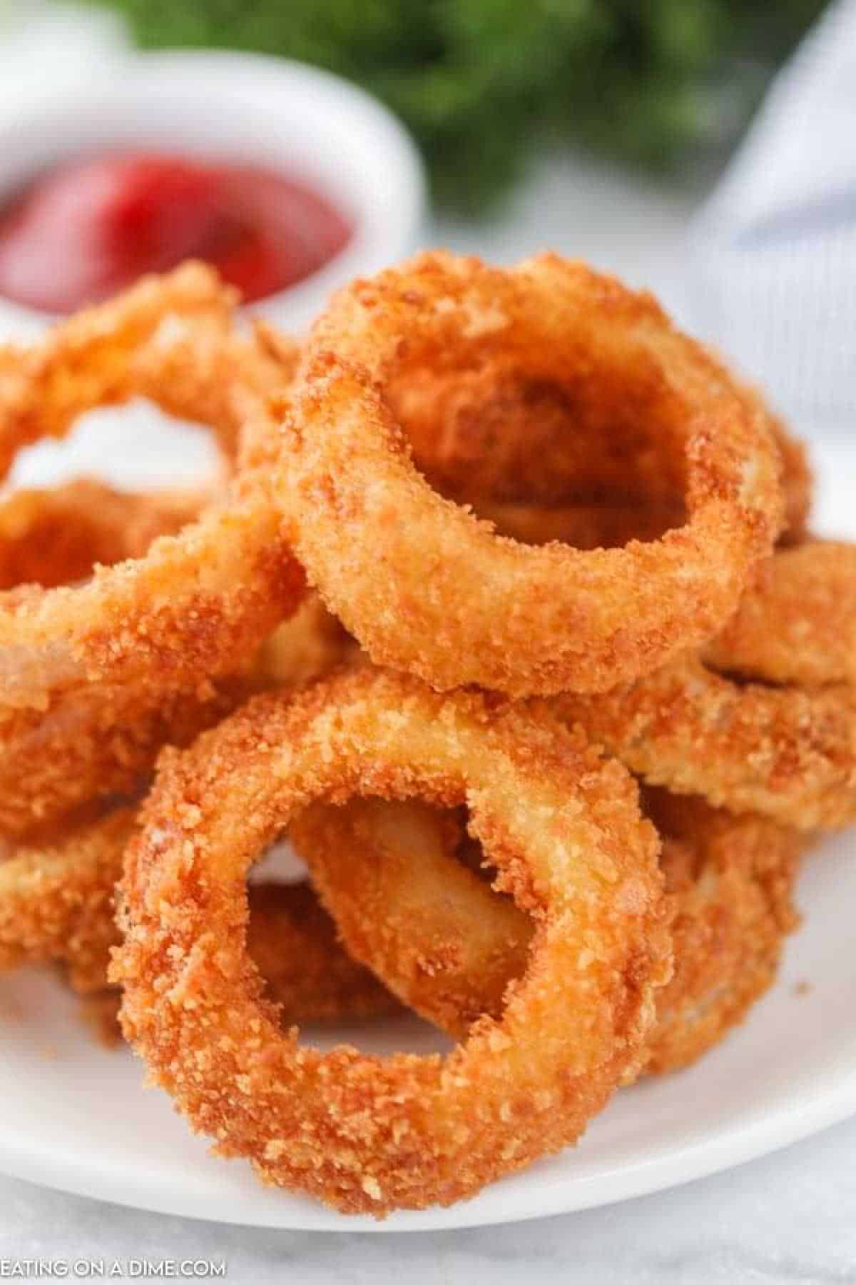 A plate of golden, crispy fried onion rings stacked high with a small bowl of red dipping sauce in the background.