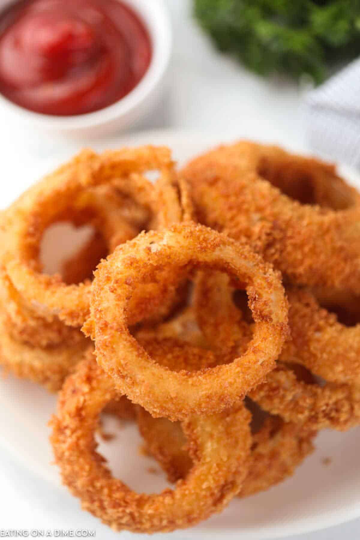 A plate of crispy, golden fried onion rings stacked high, with a small bowl of ketchup in the background and a sprig of parsley to the side.