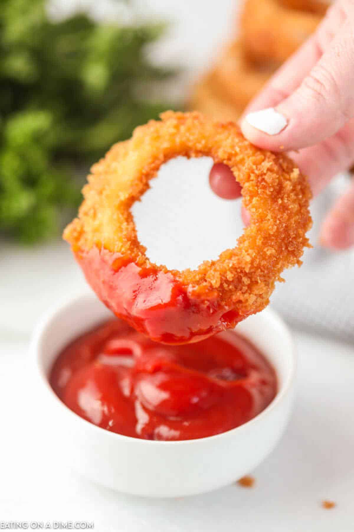 A hand holds a perfectly fried onion ring being dipped into a bowl of ketchup, with fresh green herbs in the background.