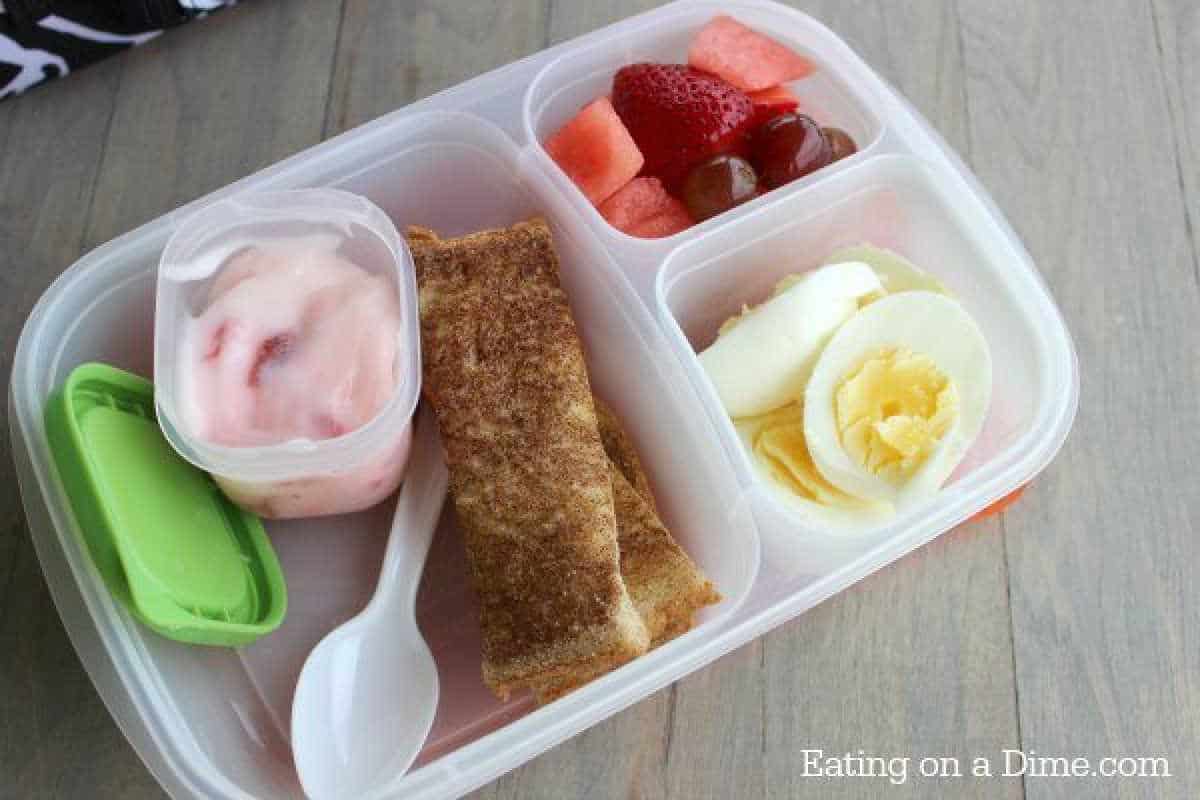 A lunchbox brimming with hard-boiled egg lunch ideas features yogurt, a green container, a cinnamon roll, a plastic spoon, fresh sliced strawberries, juicy grapes, crisp watermelon, and a perfectly sliced boiled egg nestled in separate compartments.