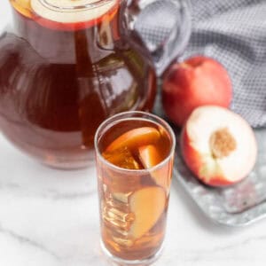 A glass pitcher and a tall glass filled with refreshing peach iced tea sit on a marble surface. The glass is garnished with peach slices. Nearby, a whole peach and a halved one rest on a metal tray with a checkered cloth, completing this delightful summer recipe scene.