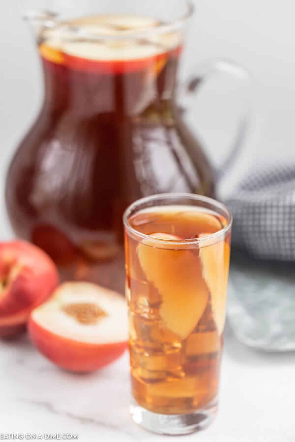 Close up image of a glass of tea with peaches in it. Also a pitcher of tea with peaches on the side. 