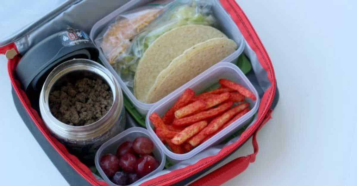 Discover how to pack tacos for lunch with this open lunchbox featuring a thermos of seasoned ground beef, crunchy taco shells, shredded cheese, juicy grapes, and a container of red corn snacks—perfect for a tasty midday treat.