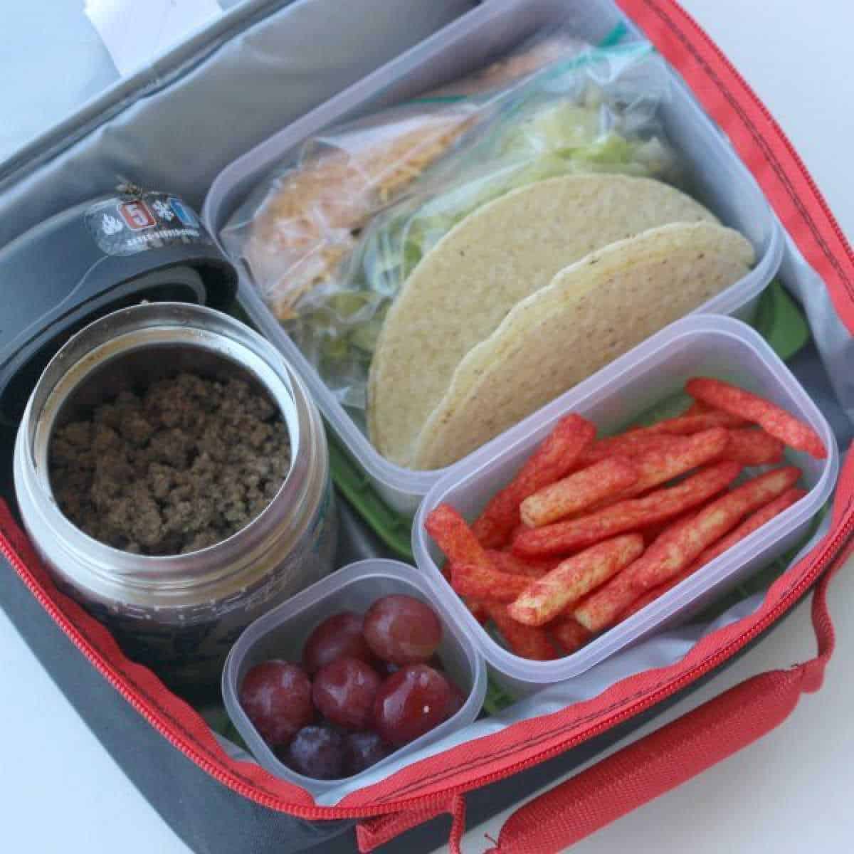 An open lunchbox reveals how to pack tacos for lunch, featuring a container of ground meat, crispy taco shells, shredded cheese, lettuce, savory red snack sticks, and juicy grapes.