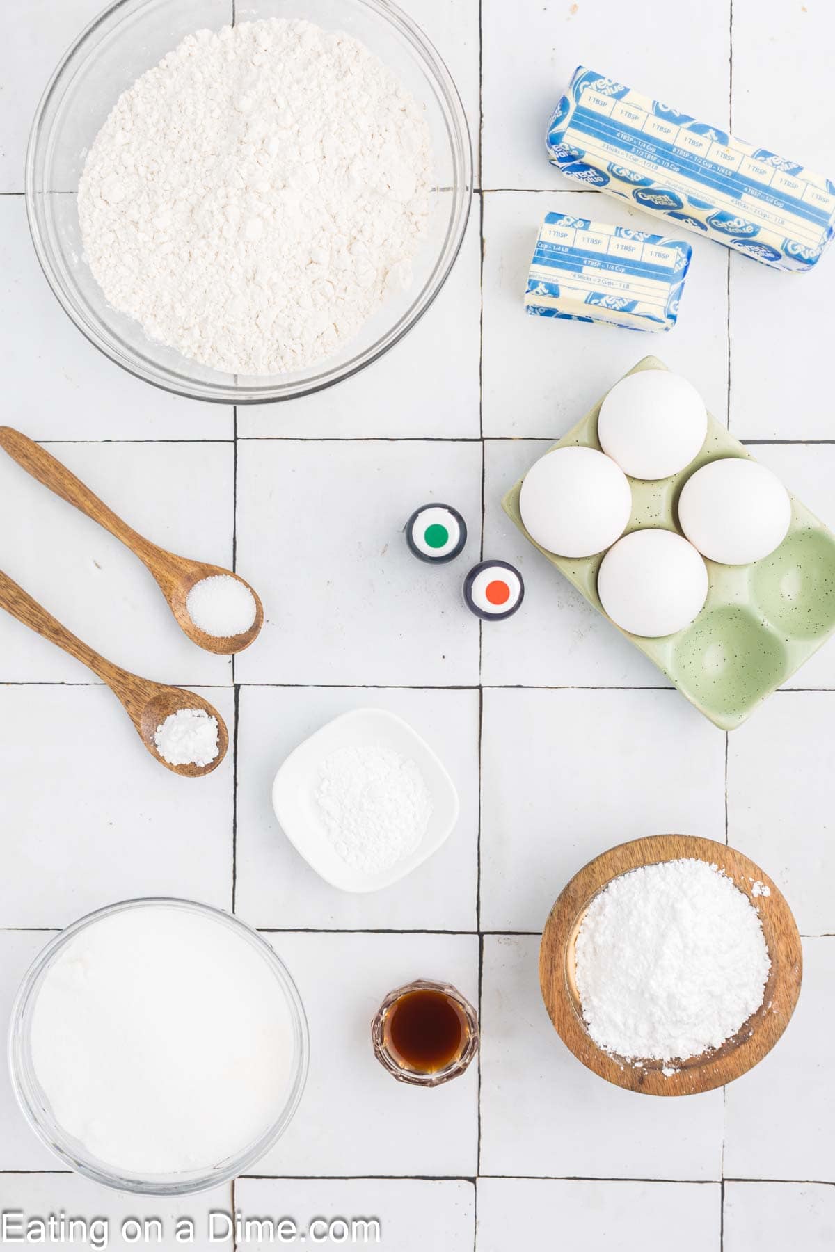 Top-down view of baking ingredients on a white tiled surface for Pumpkin Sugar Cookies. Ingredients include flour in a large bowl, four sticks of butter, four eggs in a container, two food coloring bottles, a wooden bowl of powdered sugar, a small bowl of vanilla, and bowls with salt and baking powder.