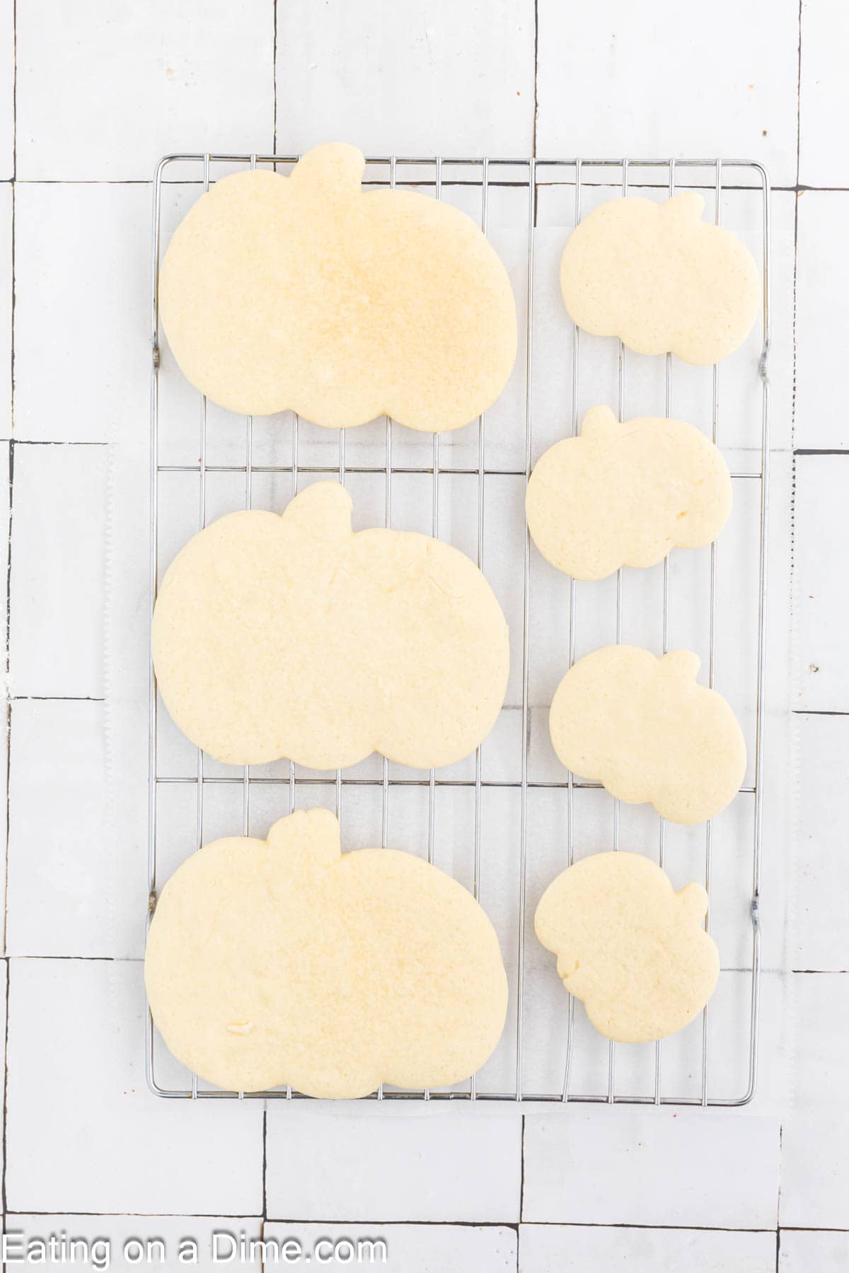 Pumpkin Sugar Cookies are cooling on a metal rack against a tiled white background. The cookies, shaped like pumpkins, are in various sizes, arranged from largest to smallest. The words "Eating on a Dime.com" are visible at the bottom left corner.