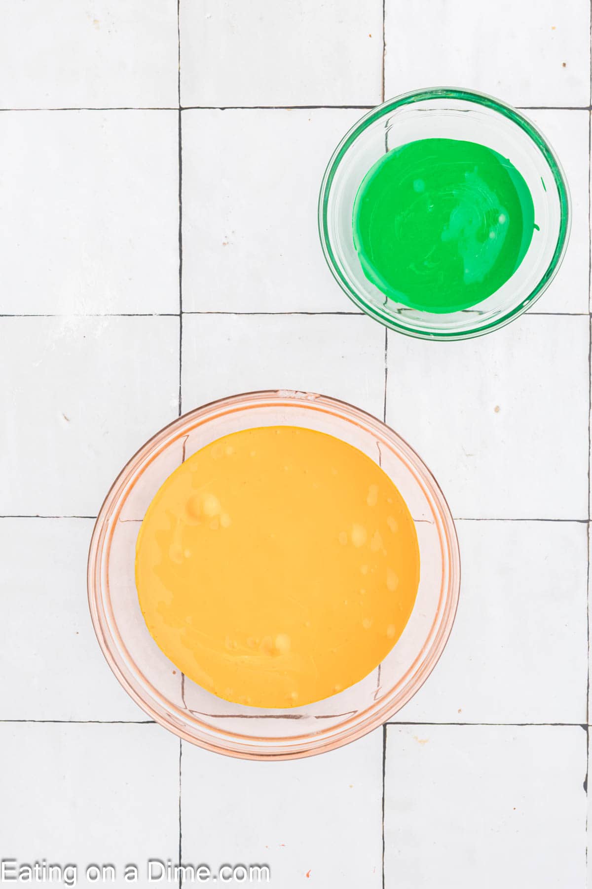 Two bowls containing icing: one with vibrant yellow icing and a smaller one with bright green icing, placed on a white tiled surface, perfect for decorating Pumpkin Sugar Cookies. The website "Eating on a Dime.com" is visible at the bottom left corner of the image.