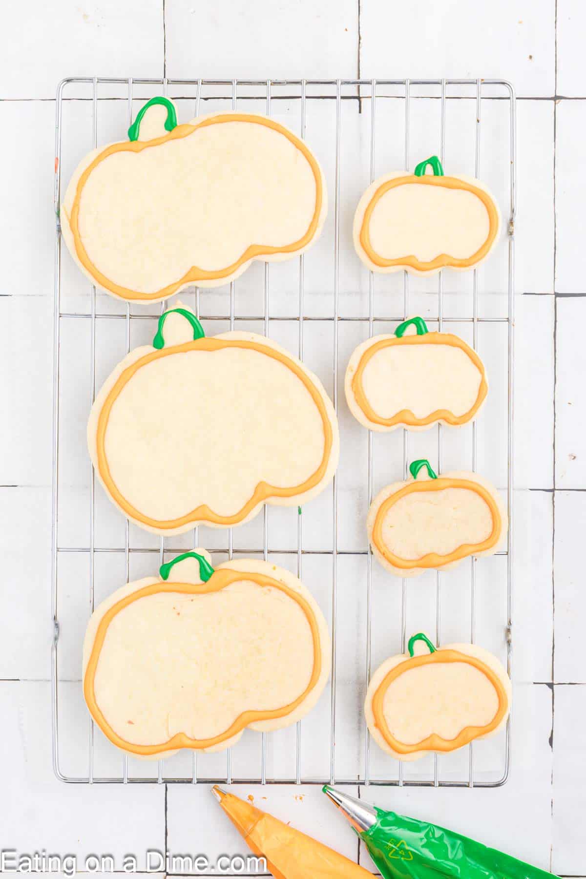 A cooling rack holds seven pumpkin-shaped sugar cookies of various sizes, decorated with simple orange and green icing details. Below, there are orange and green icing bags. The white tiled background provides the perfect backdrop for showcasing these delightful pumpkin sugar cookies.