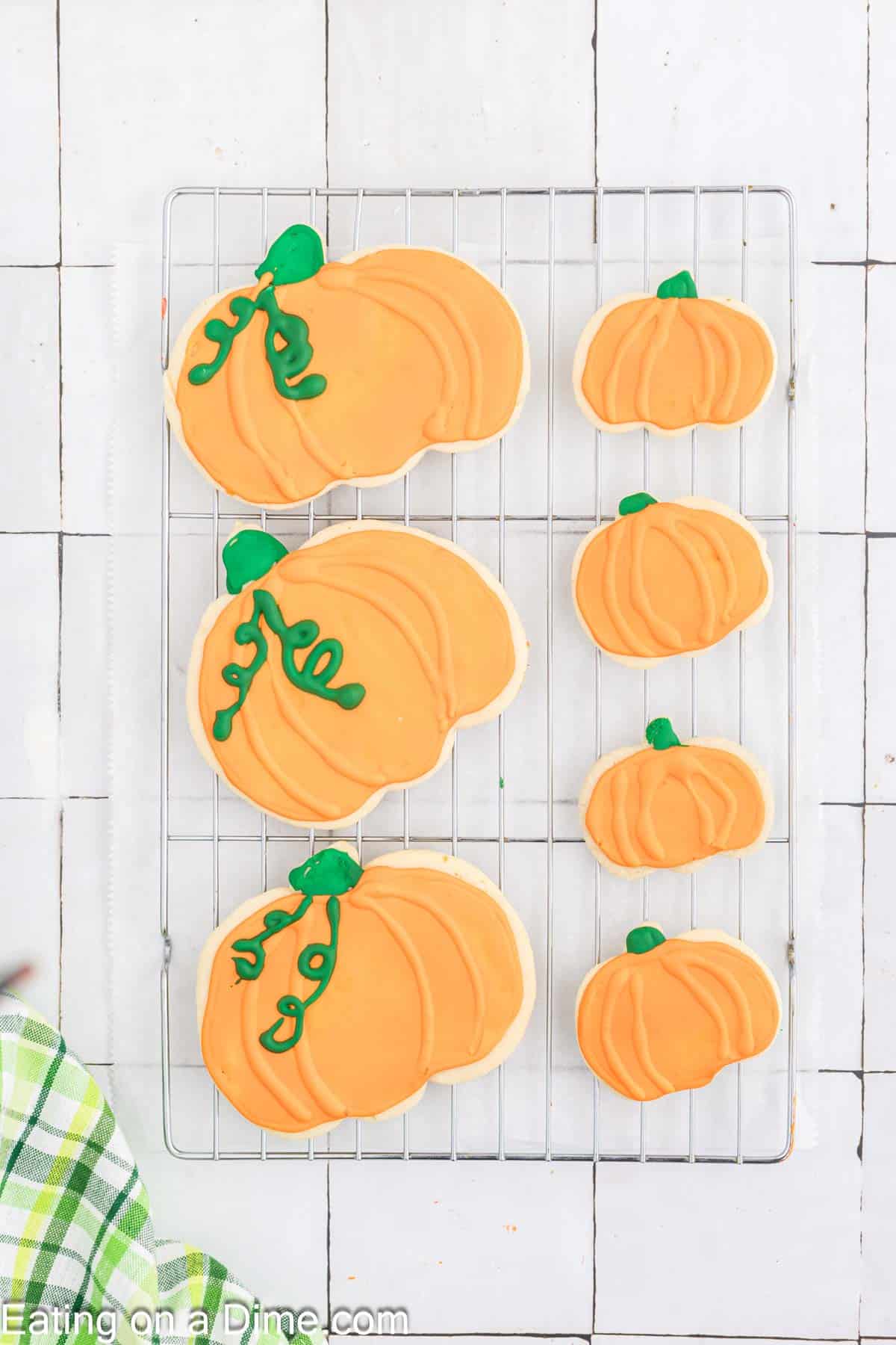 A cooling rack with six pumpkin-shaped sugar cookies is displayed on a white tiled background. The cookies are decorated with orange icing and green icing vines and leaves. A green and white kitchen towel is visible in the bottom left corner.