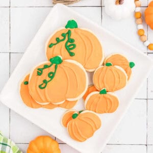 A white square plate holds eight pumpkin sugar cookies, each decorated with orange icing and green icing details for stems and vines. The plate is placed on a white tiled surface with decorative pumpkins and beads nearby.