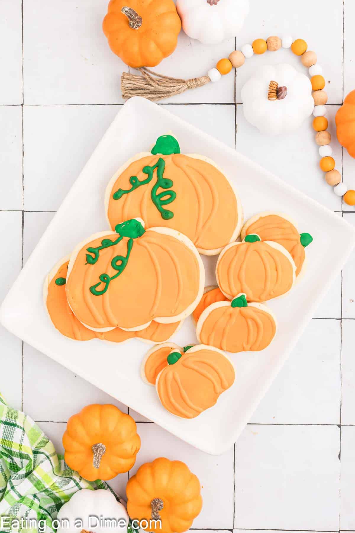 A white plate holds several pumpkin-shaped sugar cookies decorated with orange icing and green icing stems. Surrounding the plate on a white tiled surface are small decorative pumpkins and a beaded garland. A green and white checkered cloth is partially visible in the corner.