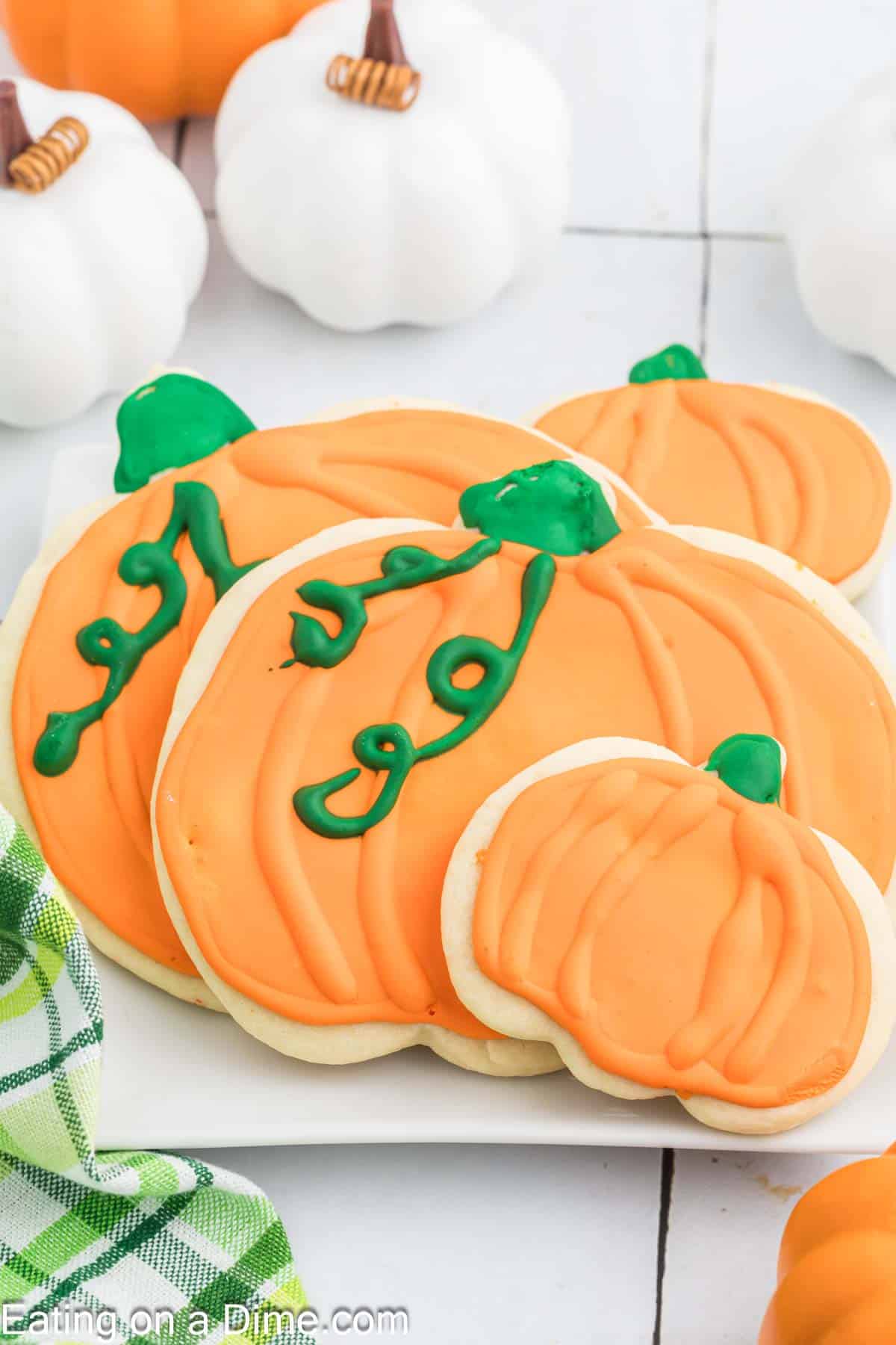 This image shows a stack of pumpkin sugar cookies with orange frosting and green icing details on a white plate. In the background, there are several white pumpkin decorations and a green-checkered cloth. The background surface is white.