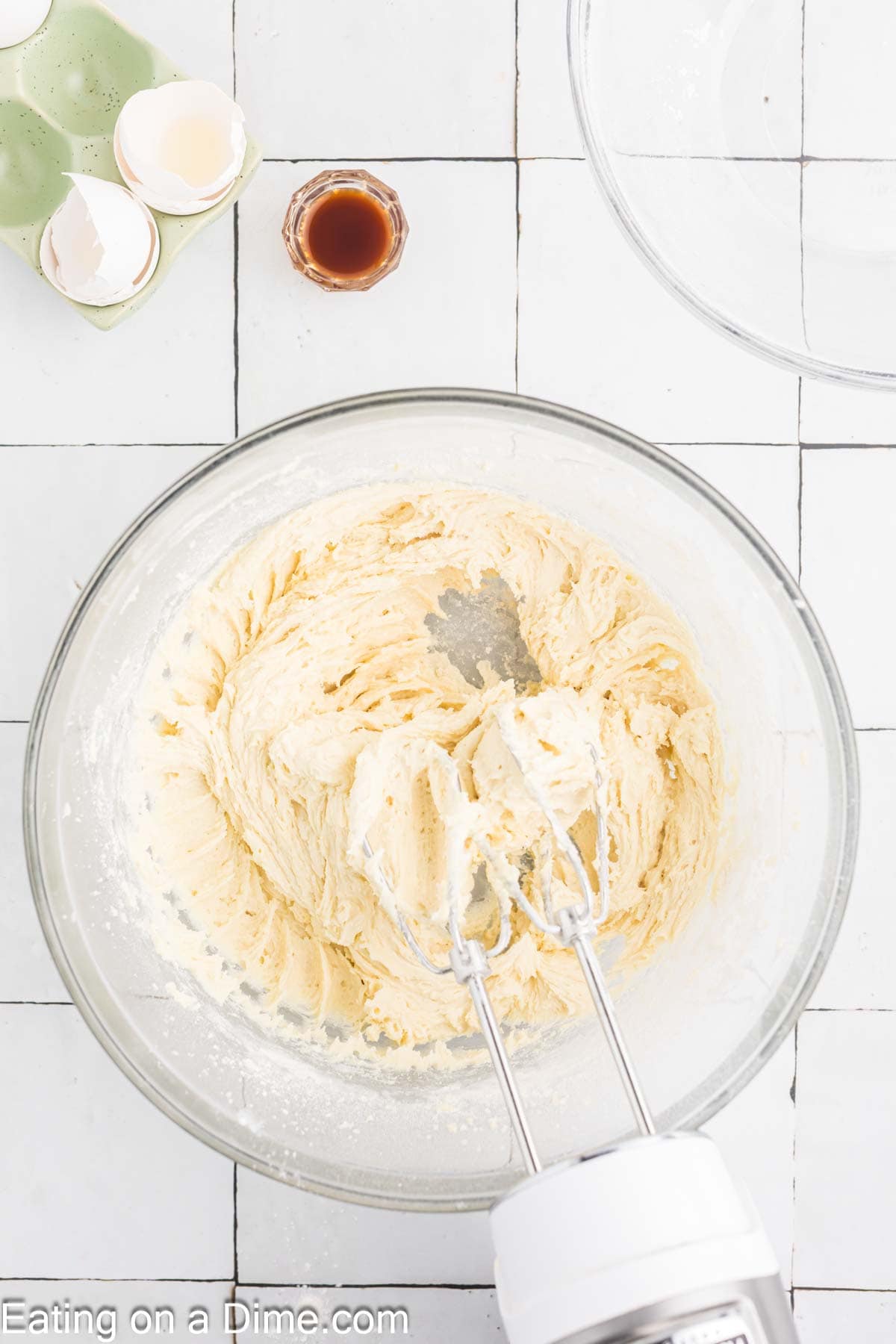 A mixing bowl filled with creamy batter for pumpkin sugar cookies and an electric mixer. Nearby are a small bottle of vanilla extract, cracked egg shells in a ceramic holder, and an empty glass bowl. The image is on a tiled surface. Text at the bottom reads "Eating on a Dime.com.