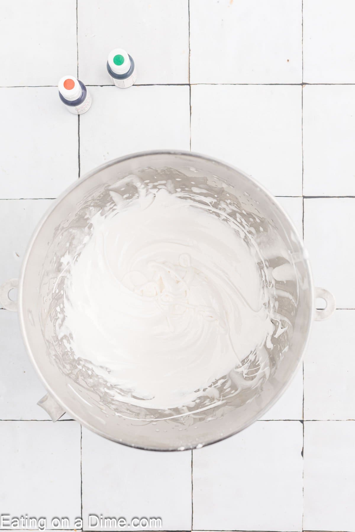 A large mixing bowl containing white frosting sits on a tiled surface. Two small bottles of food coloring, one green and one red, are placed at the top of the image. A recipe for Pumpkin Sugar Cookies can be found at the bottom, courtesy of "Eating on a Dime.com.