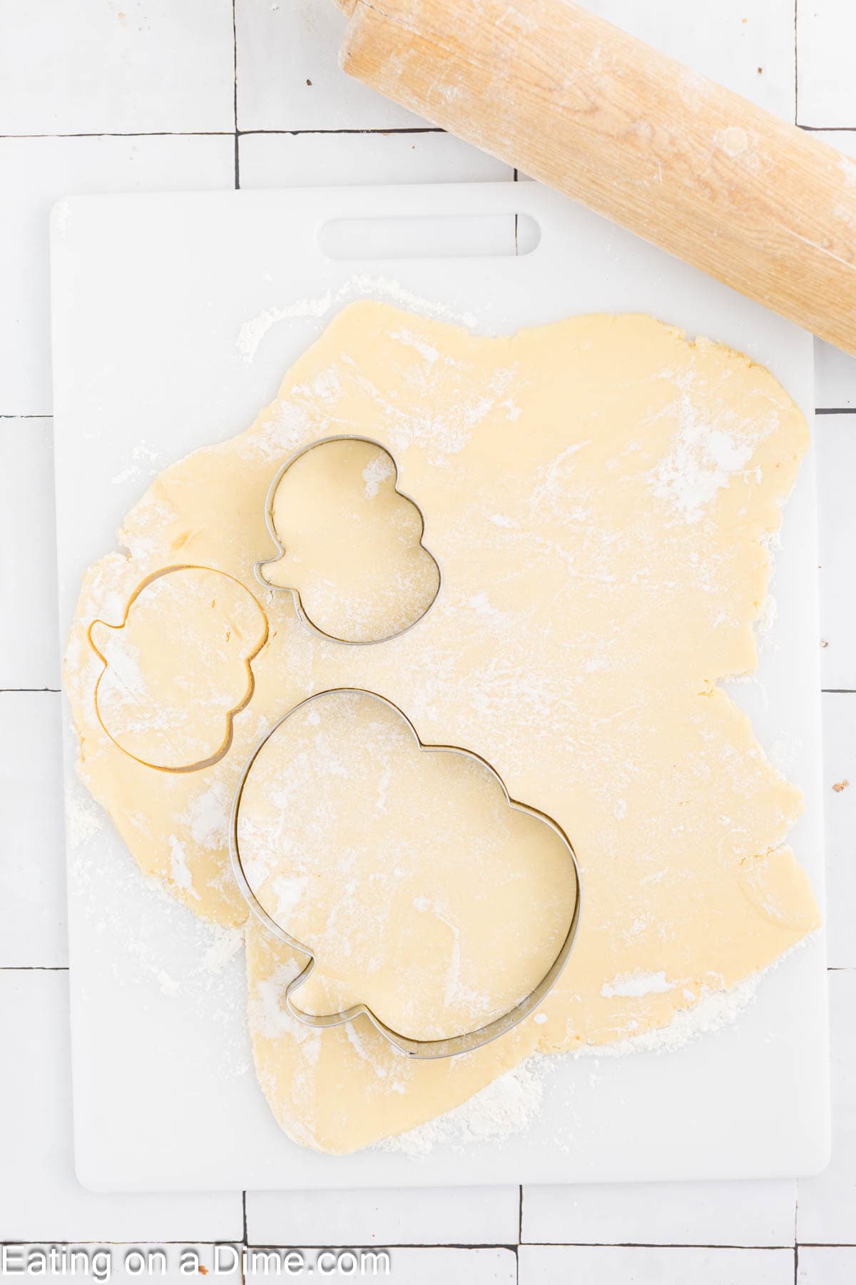 A dough on a white cutting board is rolled out flat, with excess flour scattered on top. Two metal cookie cutters in the shape of a gourd and pumpkin are placed on the dough for making Pumpkin Sugar Cookies. A wooden rolling pin rests at the top of the image.