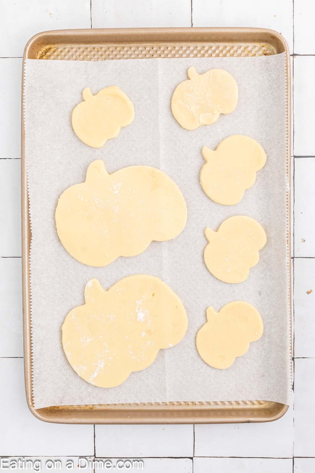 A baking tray lined with parchment paper holds several unbaked Pumpkin Sugar Cookies of various sizes. The cookies have been cut from dough and are ready to be baked. The tray rests on a white tiled surface, promising delicious treats soon.