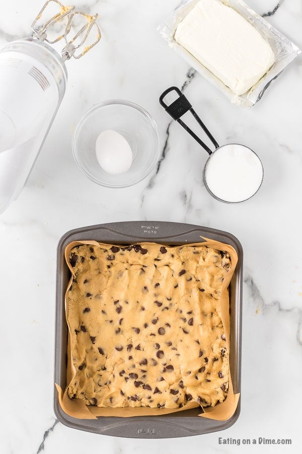 Chocolate Chip Cookie dough pressed into a 9x9 baking pan lined with parchment paper with a hand mixer, egg in a bowl, and cream cheese on this side