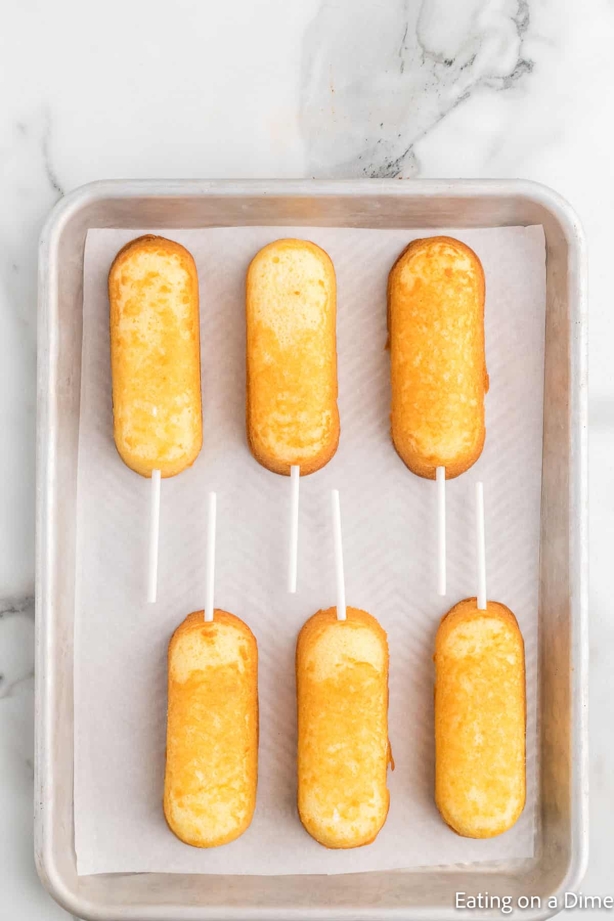 Unwrapped Twinkies with sticks in them placed on a baking sheet