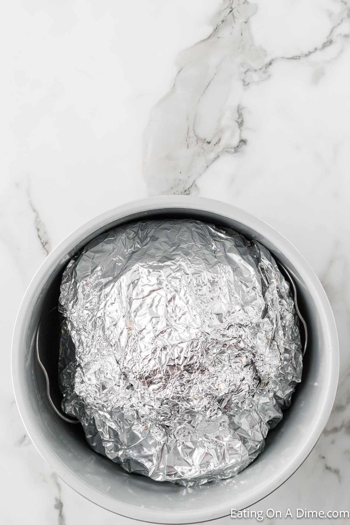 A round dish covered with aluminum foil sits inside a larger gray bowl on a marble surface. The aluminum foil is crinkled, concealing the contents entirely. The marble surface features veins in varying shades of gray. In the bottom right corner, "Eating On A Dime.com" text hints at an Instant Pot Spiral Ham Recipe within.