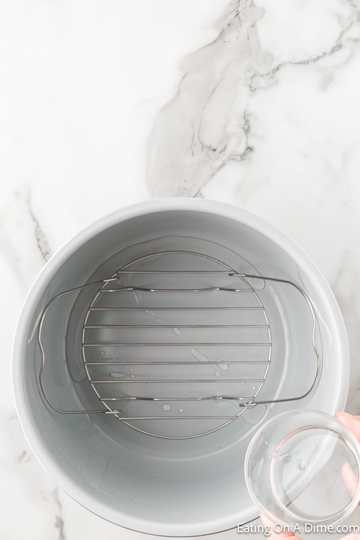 A top view of an empty pressure cooker on a white marble countertop. Water is being poured into the pot, which contains a metal rack at the bottom. The scene is well-lit and minimalistic, reminiscent of prepping for an Instant Pot spiral ham recipe, with the handle of the pouring glass partially visible.