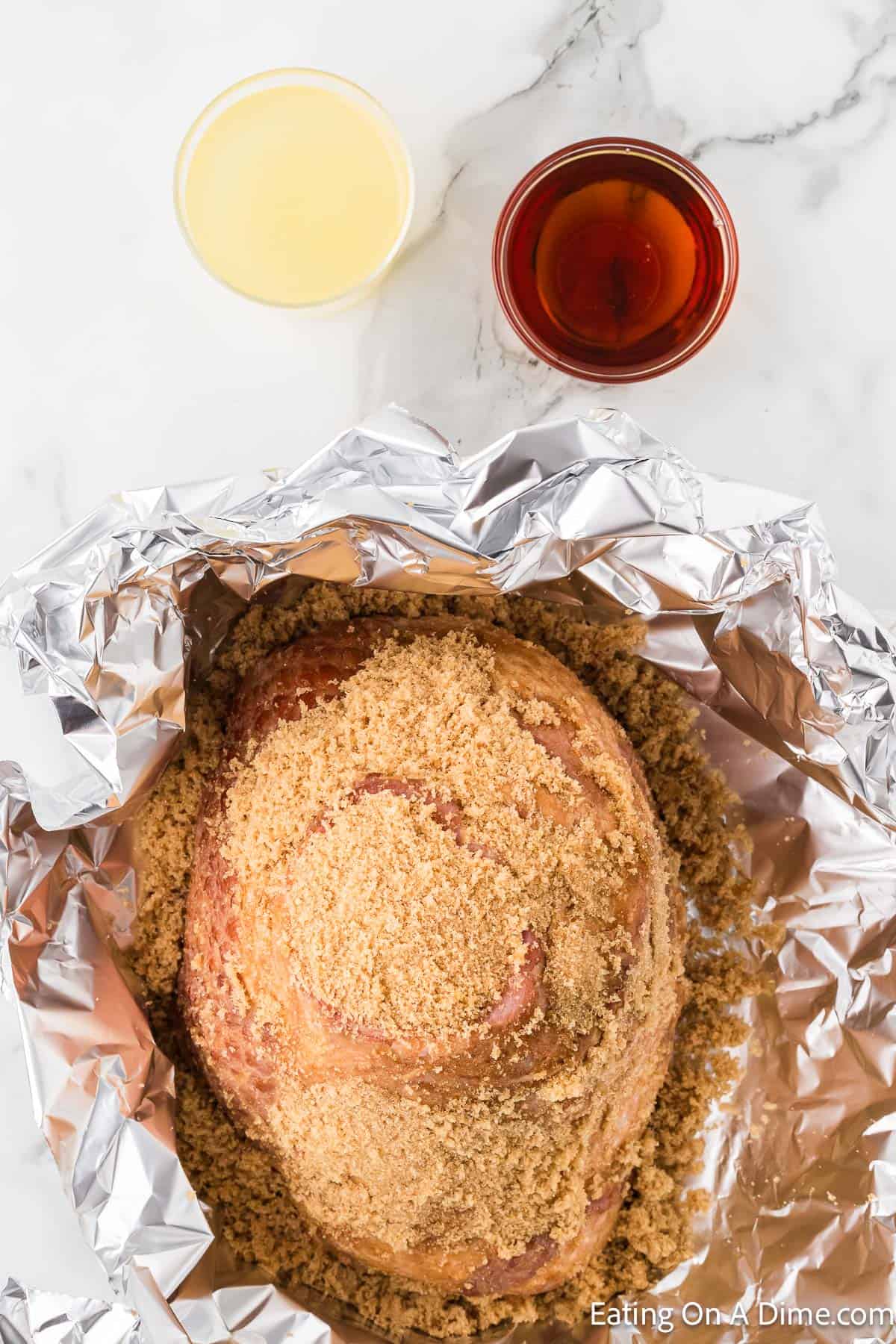 A foil-wrapped ham covered in brown sugar is prepared for baking on a marble countertop. Two small glasses of liquid, one with a yellowish drink and the other with a reddish-brown drink, are placed above the ham. The website "Eating On A Dime.com" mentions their Instant Pot Spiral Ham Recipe in the corner.