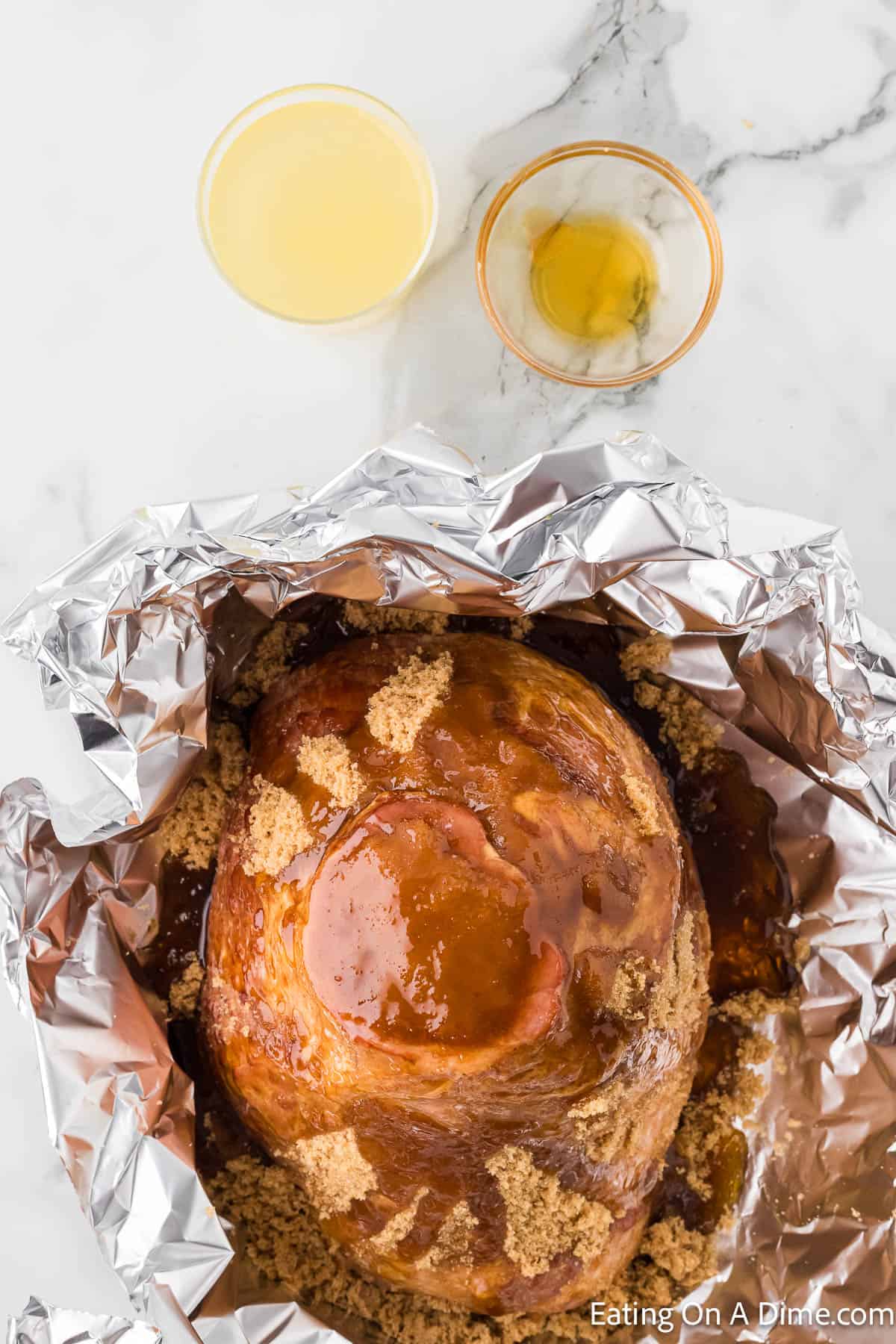 A cooked ham coated with glaze and brown sugar sits in an aluminum foil wrap on a white marble surface. Two small glasses beside the ham contain liquids, one light yellow and the other a darker amber color. This tempting dish follows an Instant Pot Spiral Ham recipe for effortless cooking.