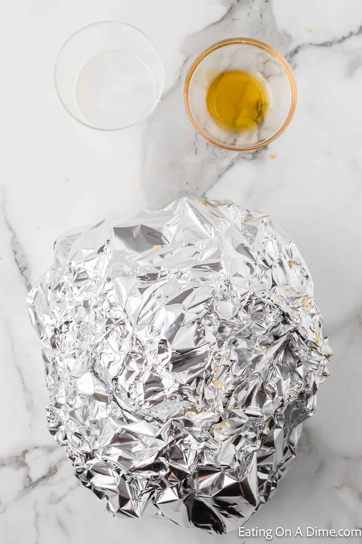 A marble countertop with two small bowls, one containing a clear liquid and the other a yellow liquid. Below the bowls is a ball of crumpled aluminum foil. The bottom right corner features an “Eating On A Dime.com” watermark, perfect for showcasing your Instant Pot Spiral Ham Recipe.